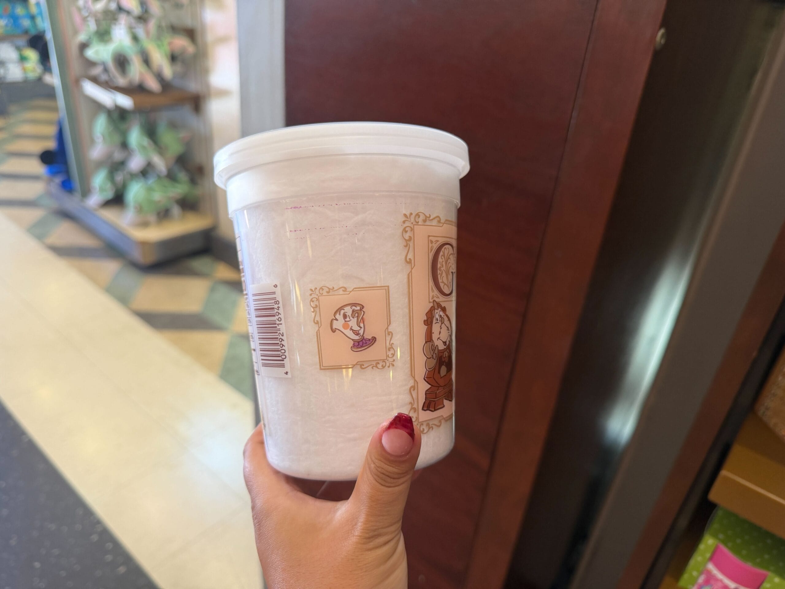 A hand with red nails holds a clear tub of white cotton candy, possibly a sweet treat from a Disney park snack shop or gift store aisle.