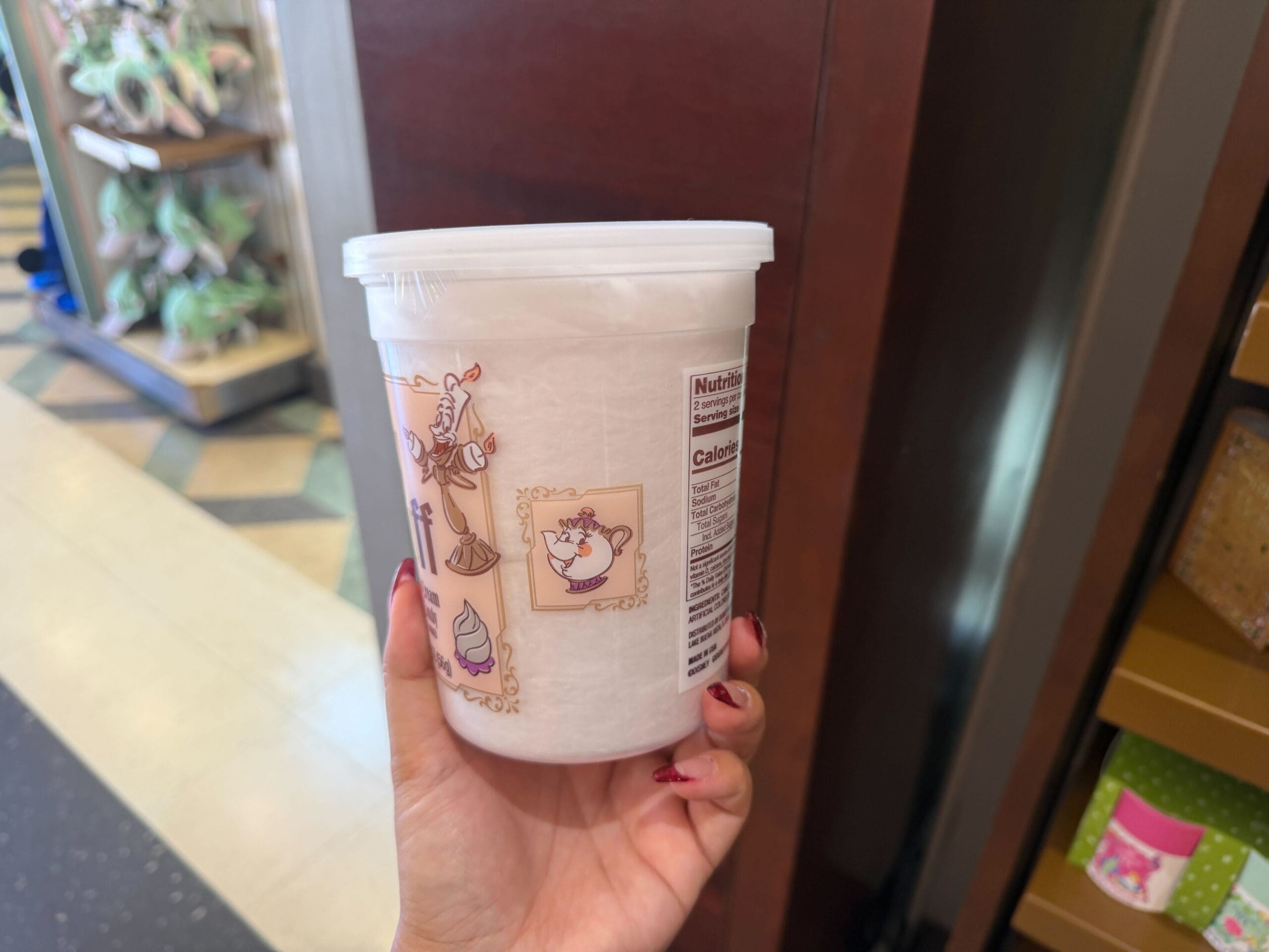 A hand holds a Beauty and the Beast cotton candy tub in a Disney park shop, label features Belle and Beast like park merchandise.