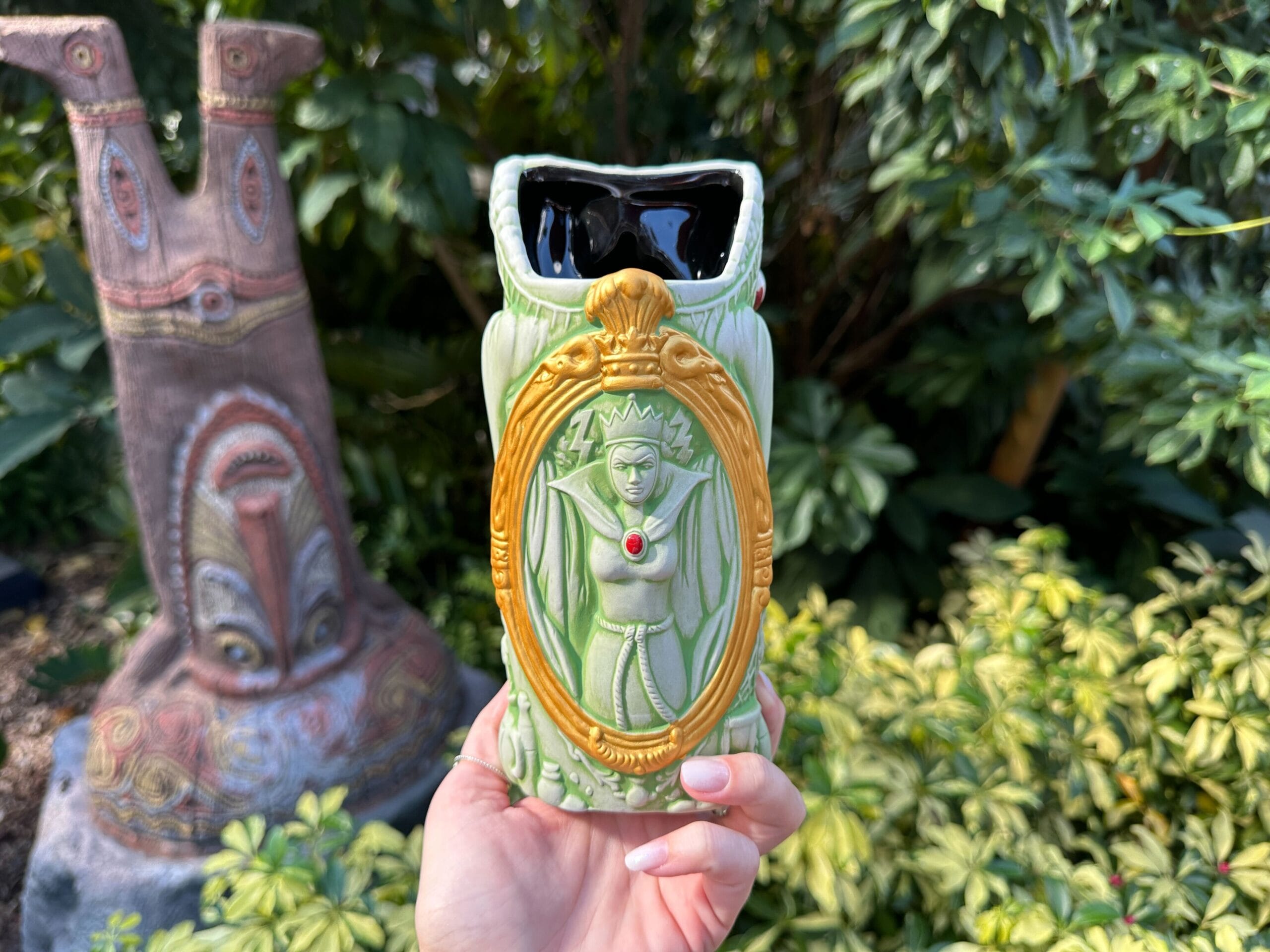 A hand displays a green Evil Queen Tiki Mug amid lush plants and a tiki statue, reminiscent of Disney's Polynesian Village Resort.