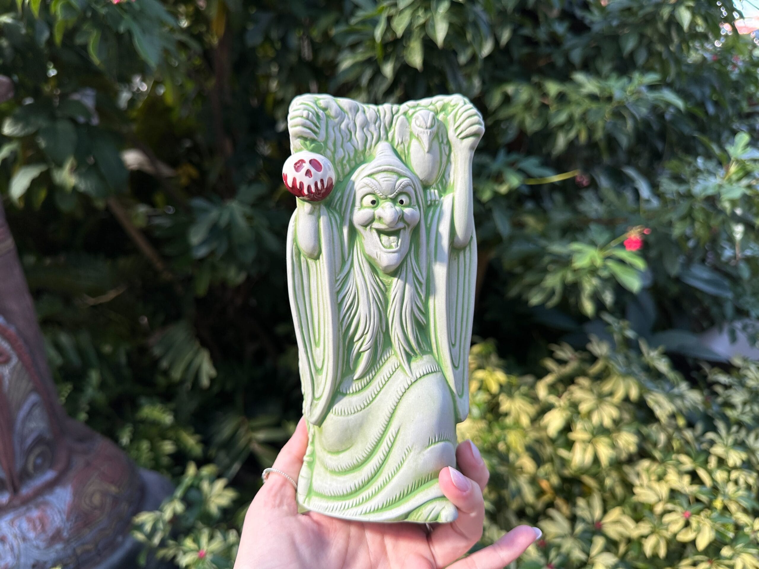 A hand holds an Evil Queen Tiki Mug, shaped like the witch with a poison apple, lush Disney park foliage behind it.