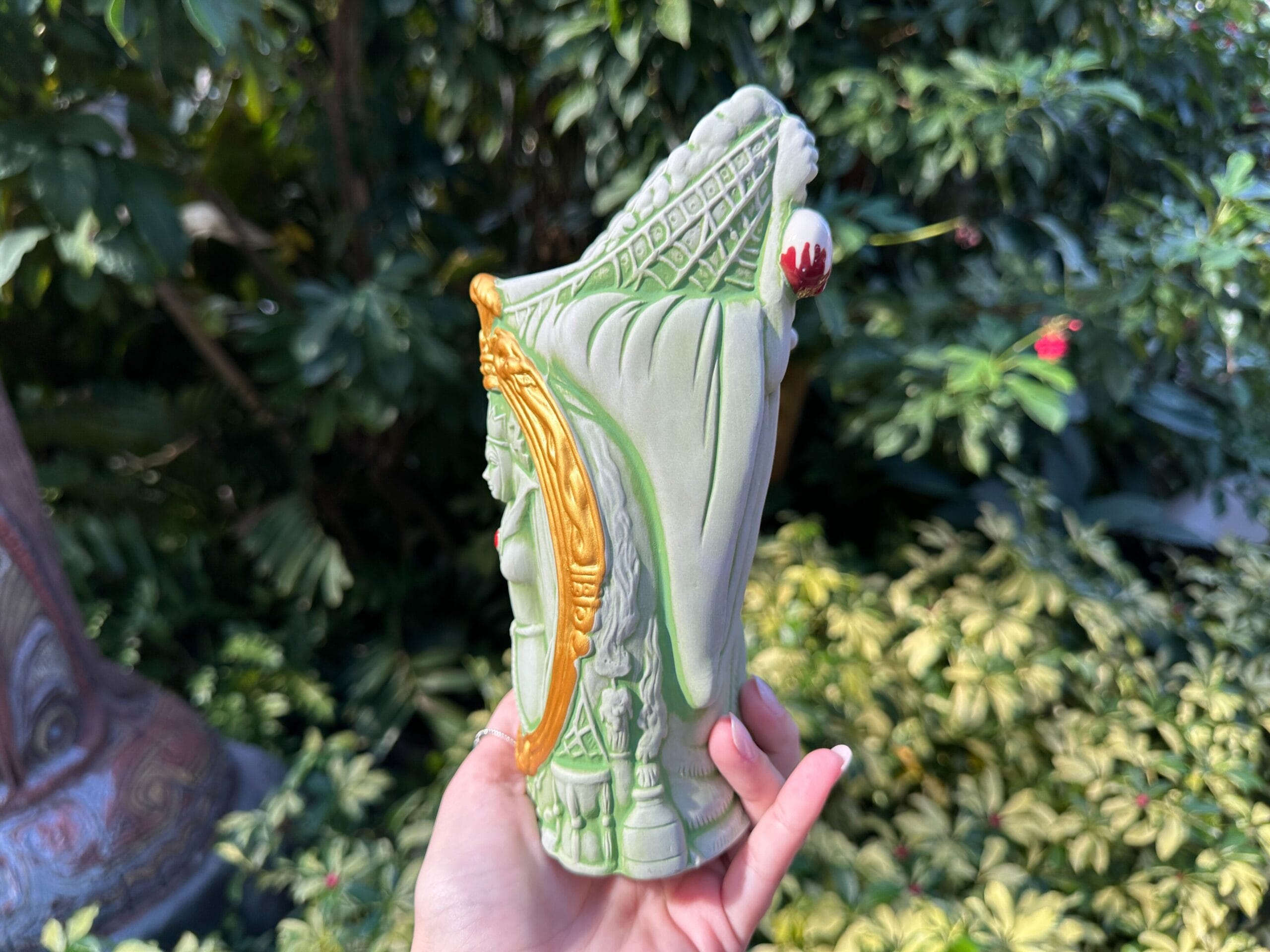 A hand holds a green Evil Queen Tiki Mug, resembling a Disney villain, with lush plants behind—no castle like Disney’s in view.