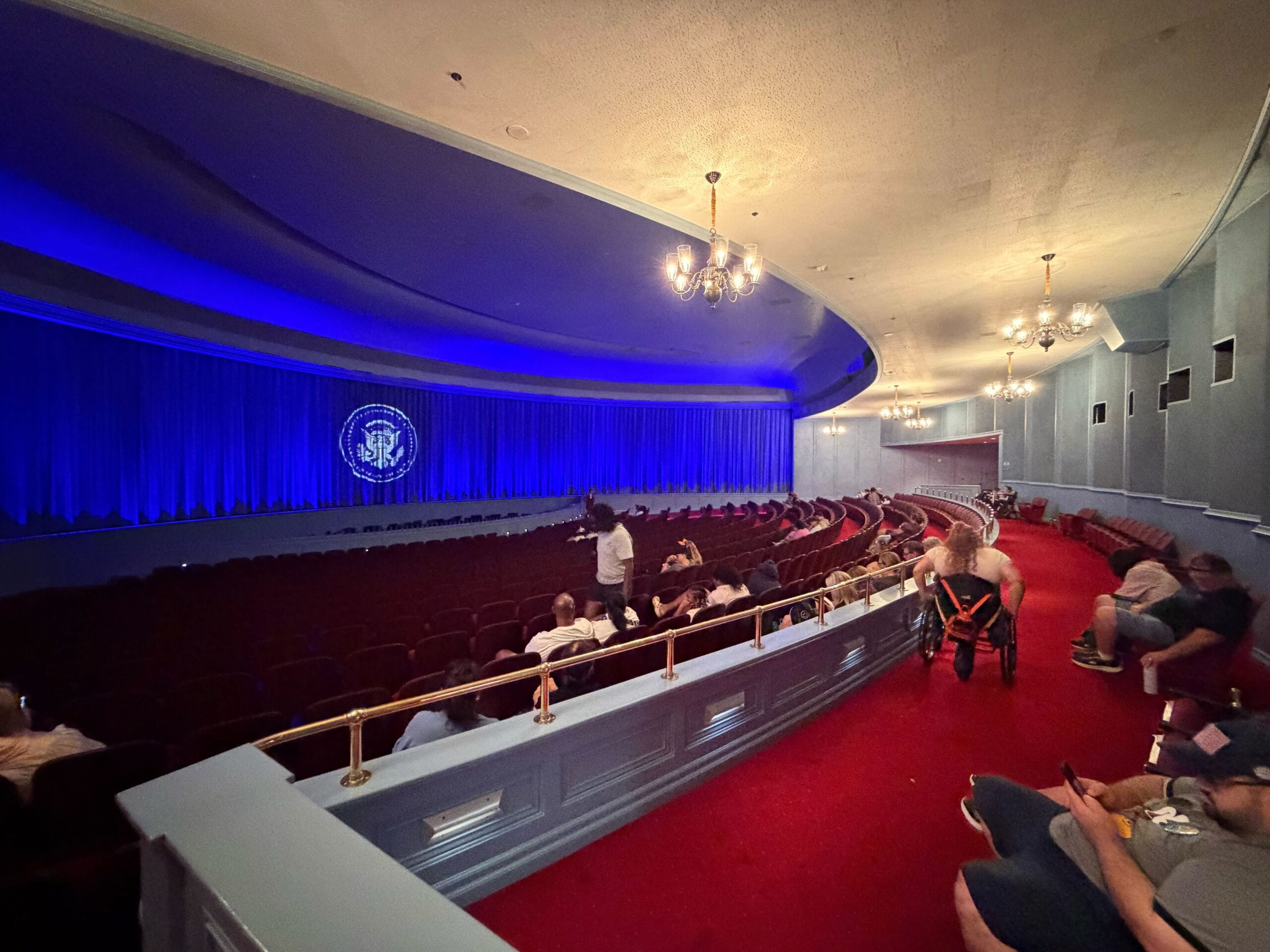 Spacious Disney theater with blue curtains, red carpet, and guests seated, waiting for entertainment to begin—no castle in view.