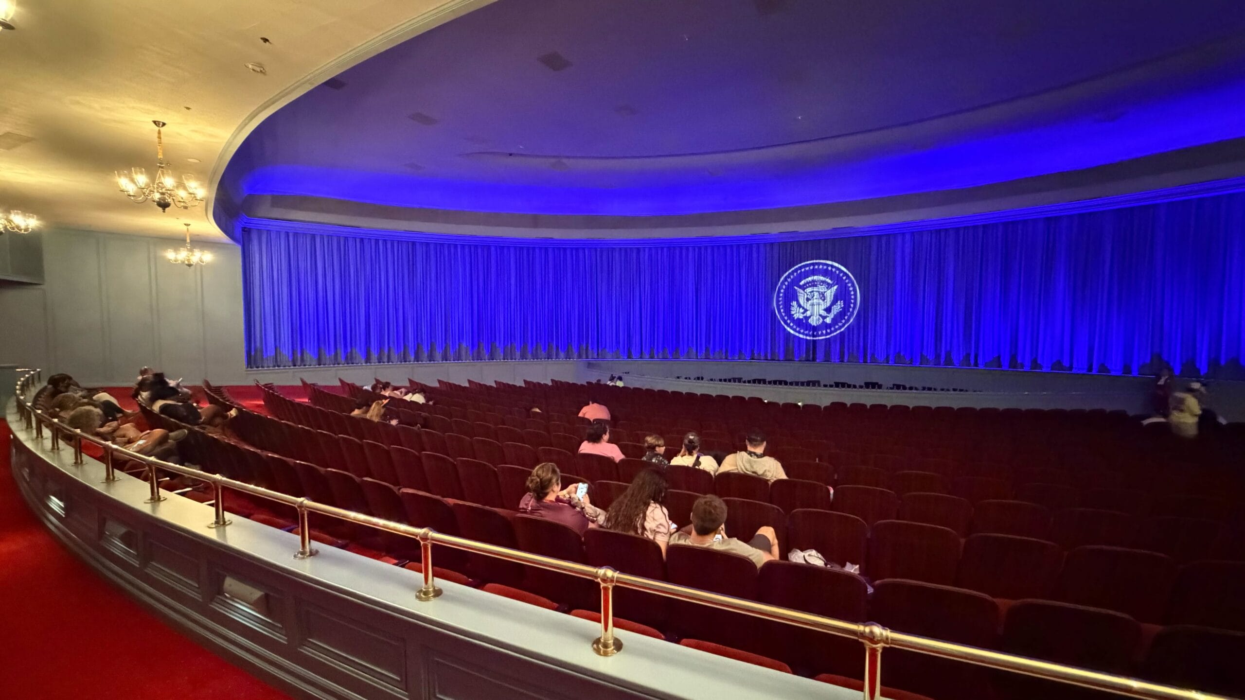 Audience in a theater resembling the Hall of Presidents at Disney Parks, blue-lit stage, U.S. presidential seal on curtains.