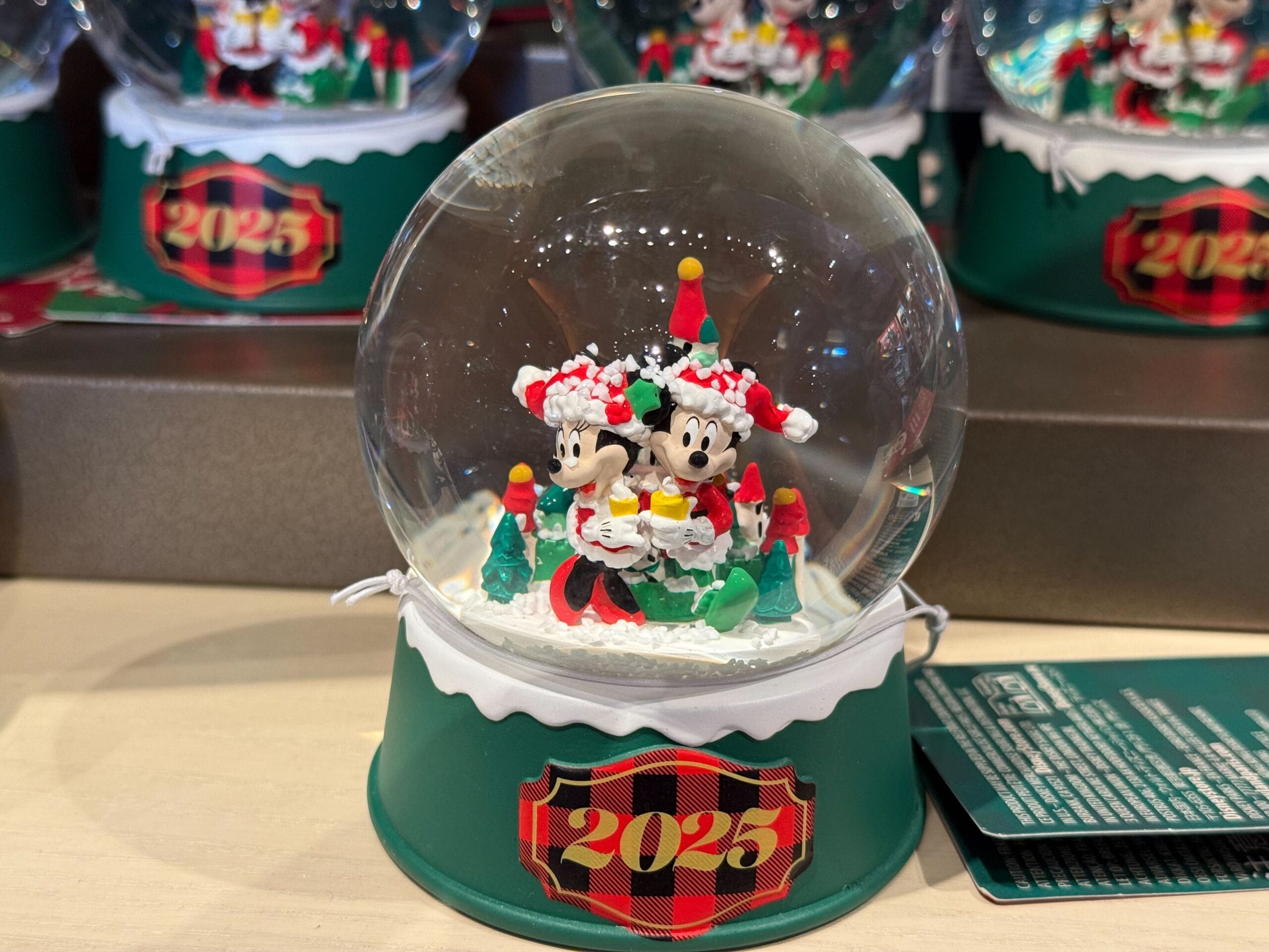 A Disneyland Resort Christmas snow globe featuring Mickey and Minnie in festive outfits, "2025" on a green base, no castle shown.