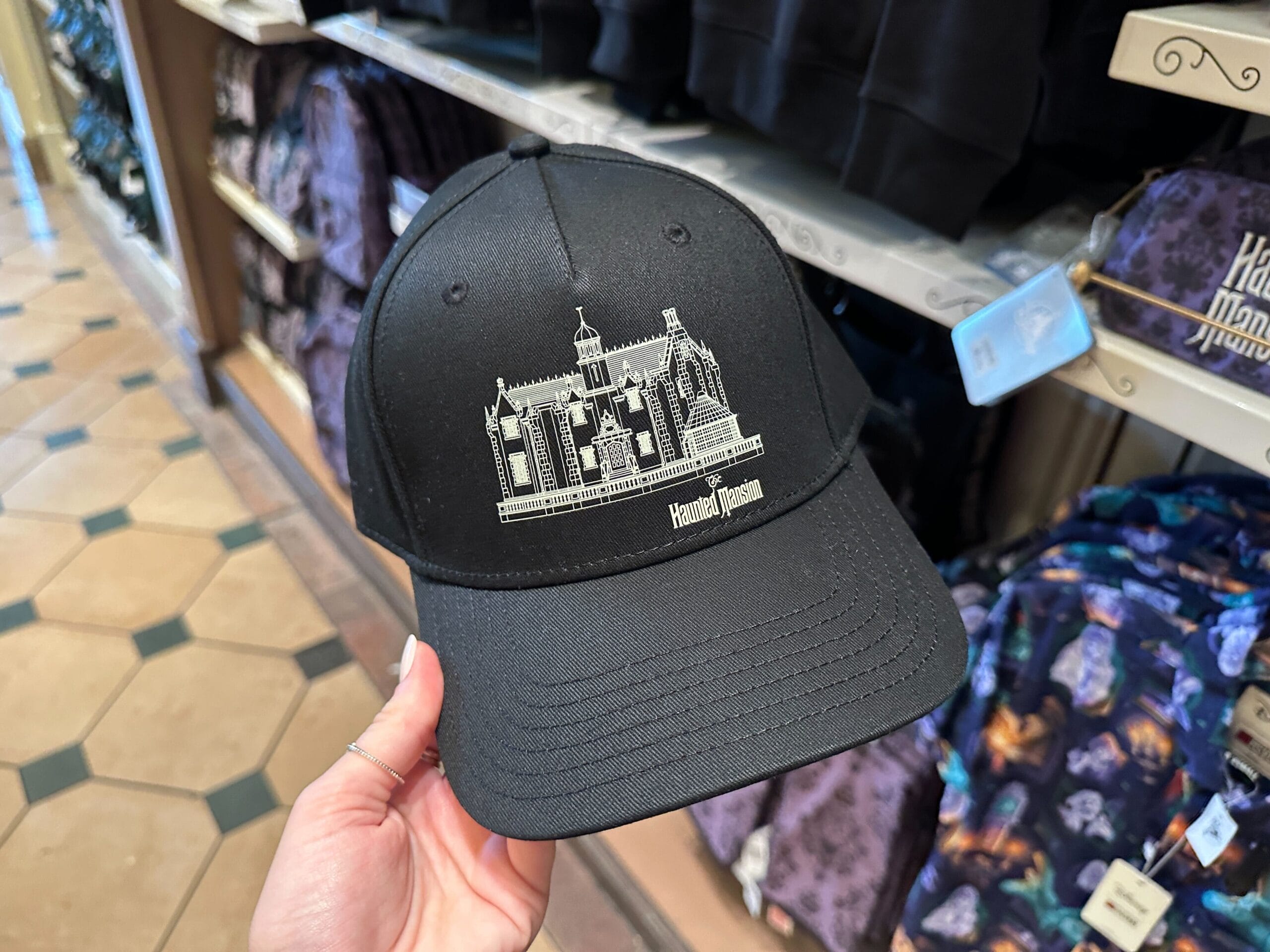 A hand holds a black Haunted Mansion hat with a white outline of the iconic mansion featured in Disney Parks around the world.