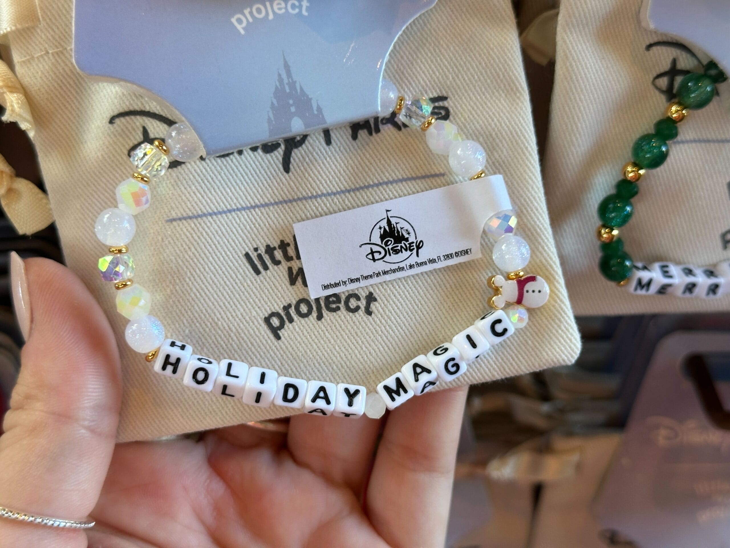 A hand holds a beaded bracelet reading “HOLIDAY MAGIC” with Disney park packaging in the background, reminiscent of park merchandise.
