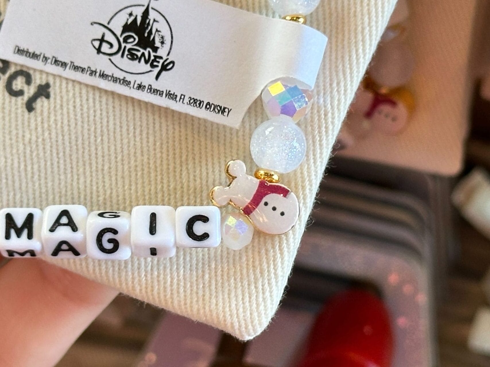 Close-up of a beaded bracelet spelling "MAGIC" with a Disney park tag and character charm, held above tan cloth, no castle visible.