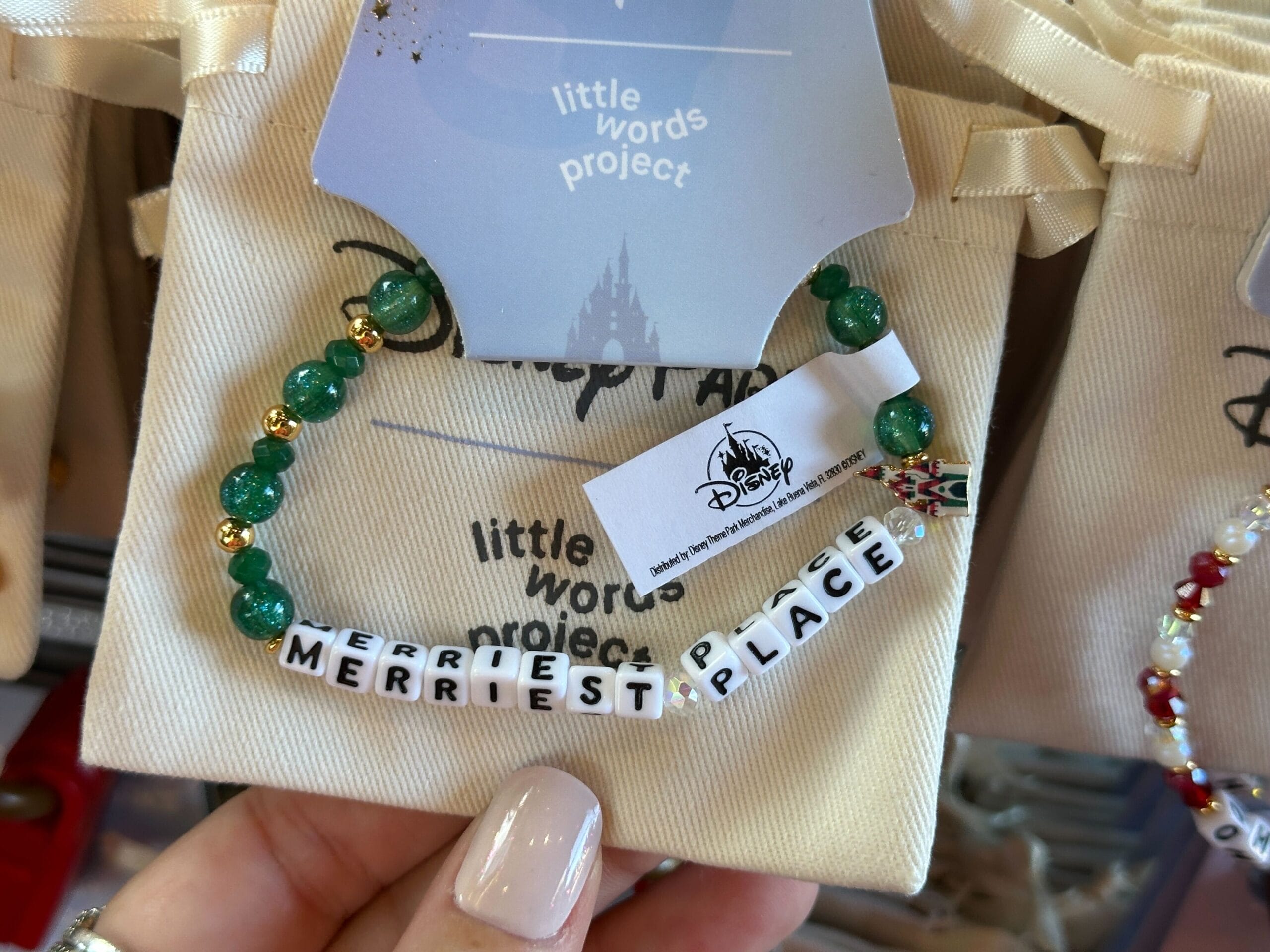 A beaded bracelet with white cube beads spelling "MERRIEST PLACE" displayed on a Disney-themed card, evoking park festivities.