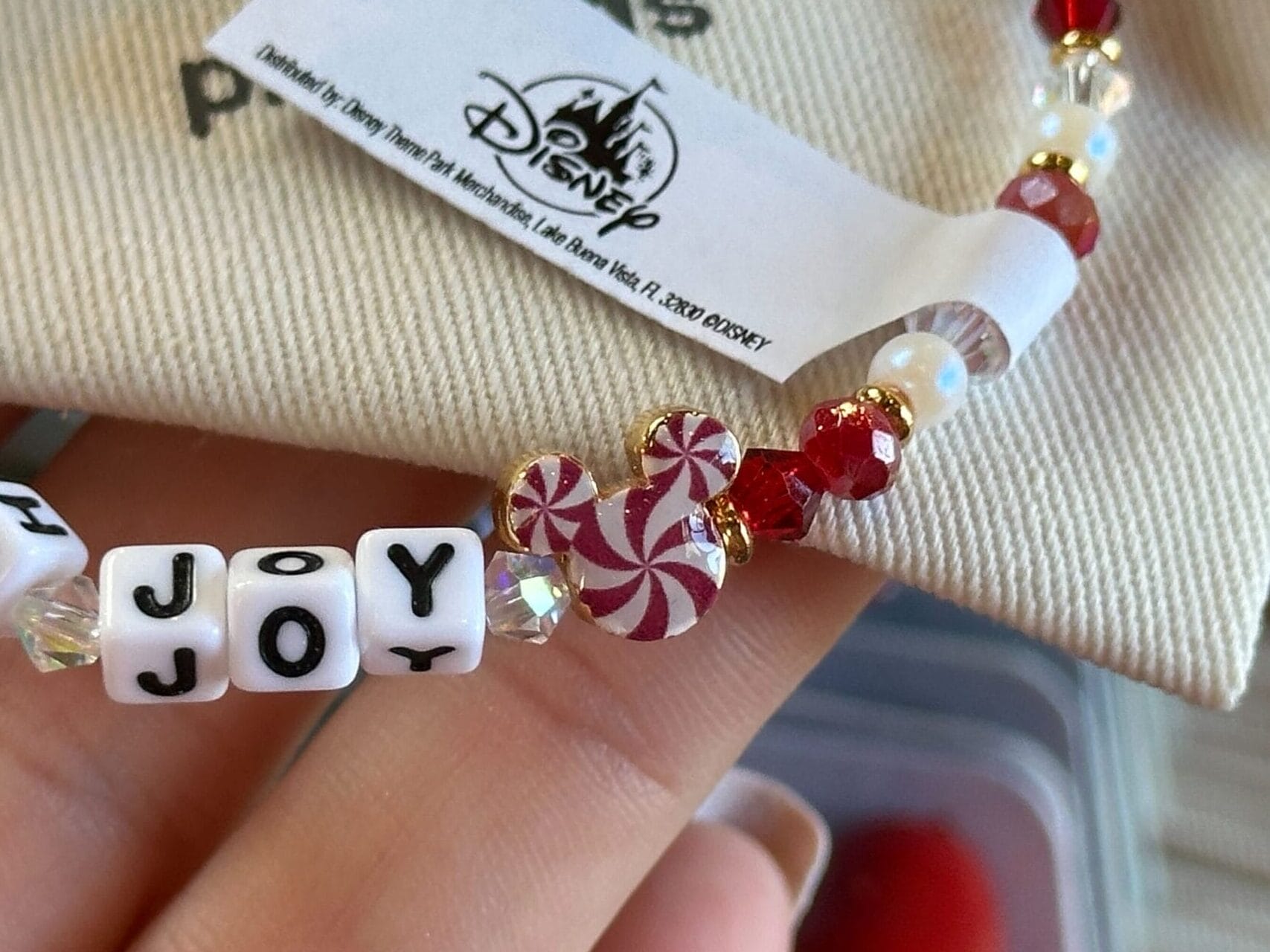 Beaded bracelet with red and white beads and "OH JOY" letter blocks, held up in front of a Disneyland tag, reminiscent of park souvenirs.