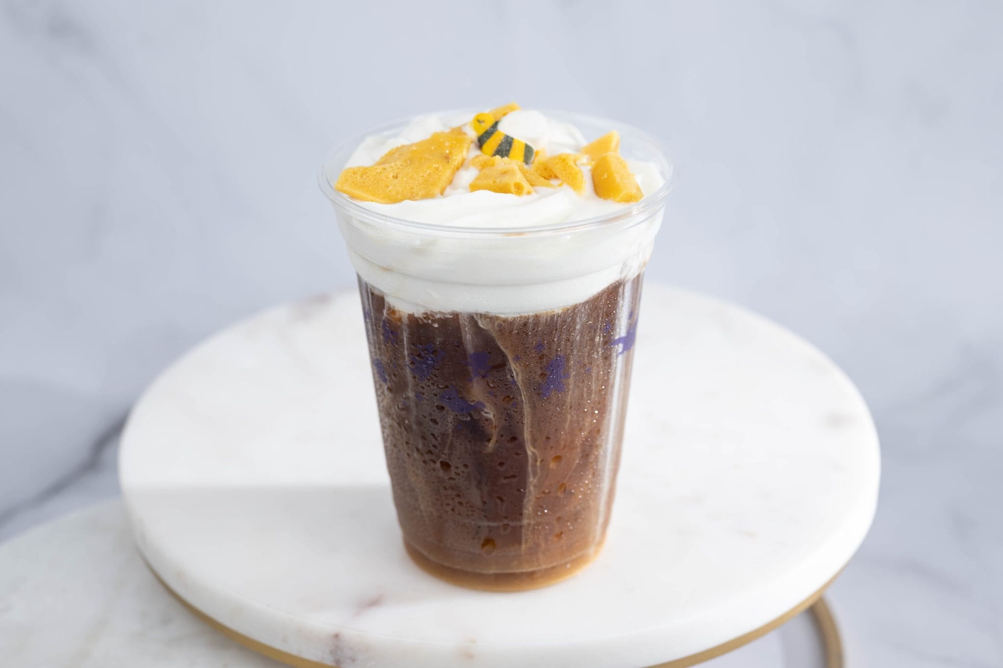 Cold Brew topped with whipped cream and yellow fruit pieces in a clear plastic cup on a white marble surface.