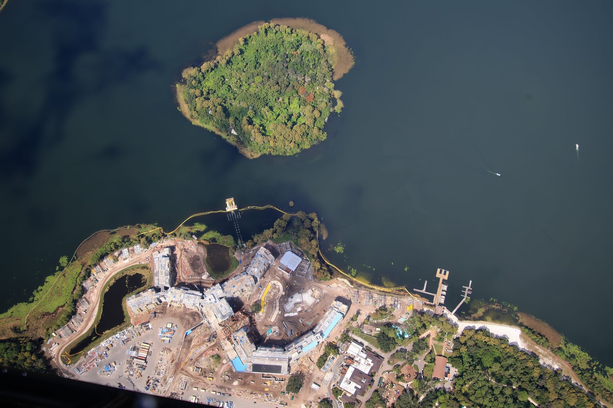 Aerial view of Disney Lakeshore Lodge construction site by a lake with docks and a small, green island, similar to the scenery near Disney parks.