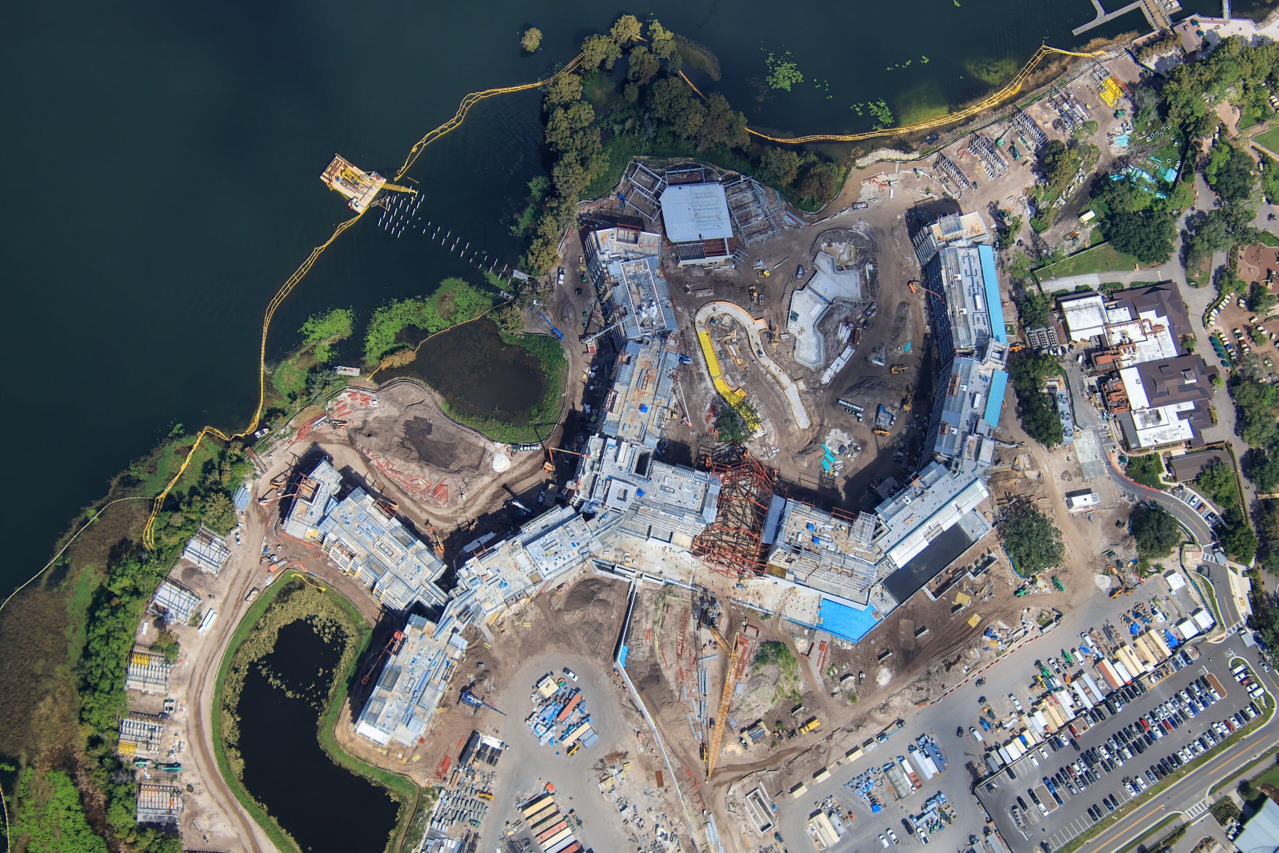 Aerial view of a large building under construction near a lake at a Disney park, with roads and parked vehicles, no castle visible.