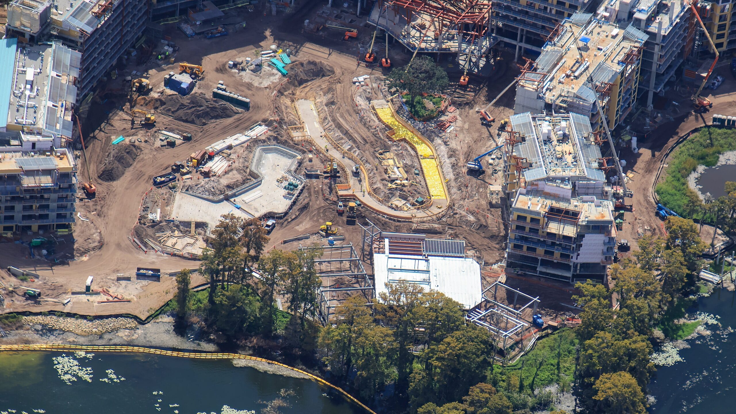 Aerial view of Disney Lakeshore Lodge construction site near water, surrounded by buildings and trees, similar to expansion areas seen at Disney parks.