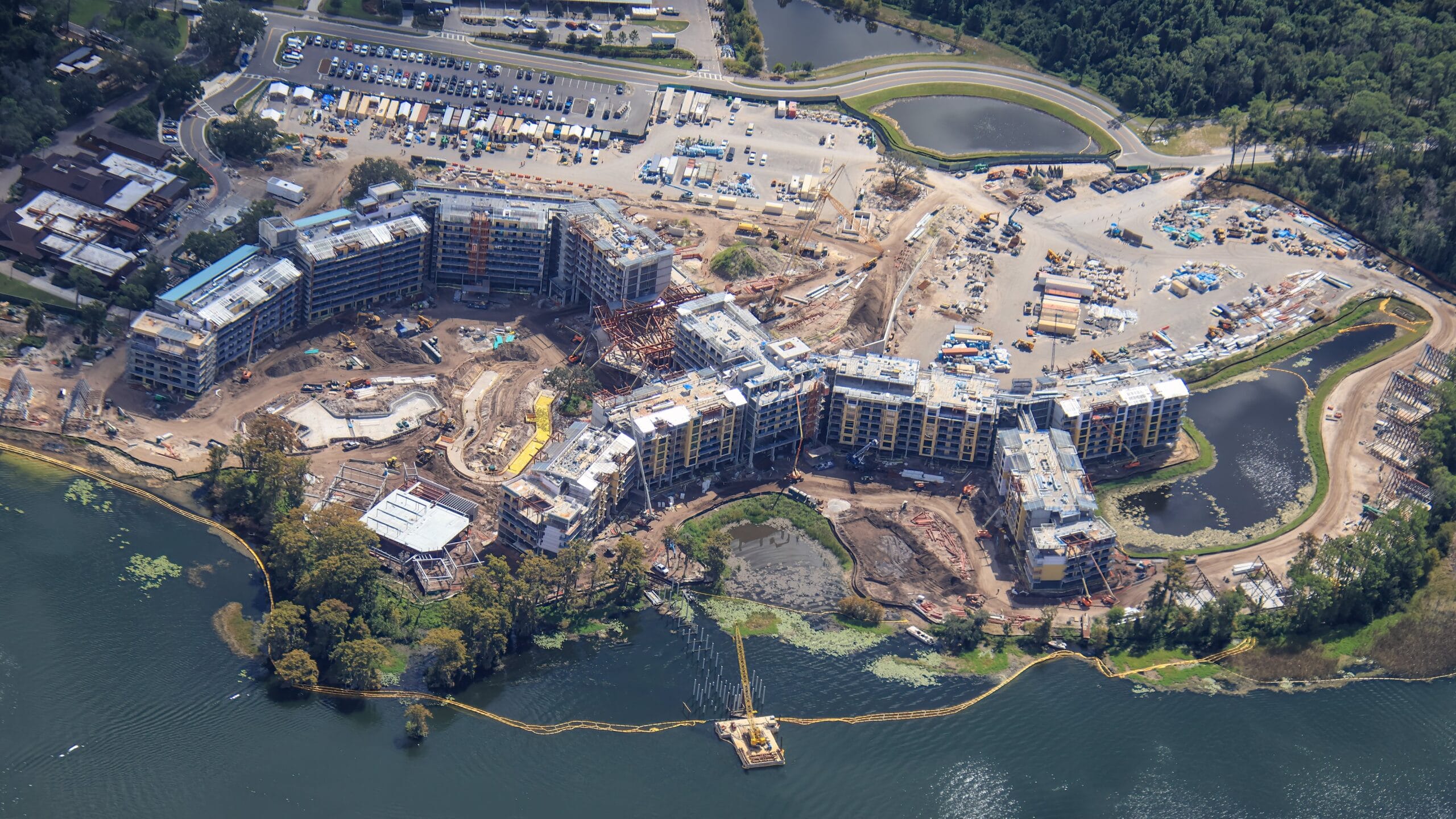 Aerial view of a large lakeside construction site with multiple buildings and cranes, reminiscent of Disney park expansions.