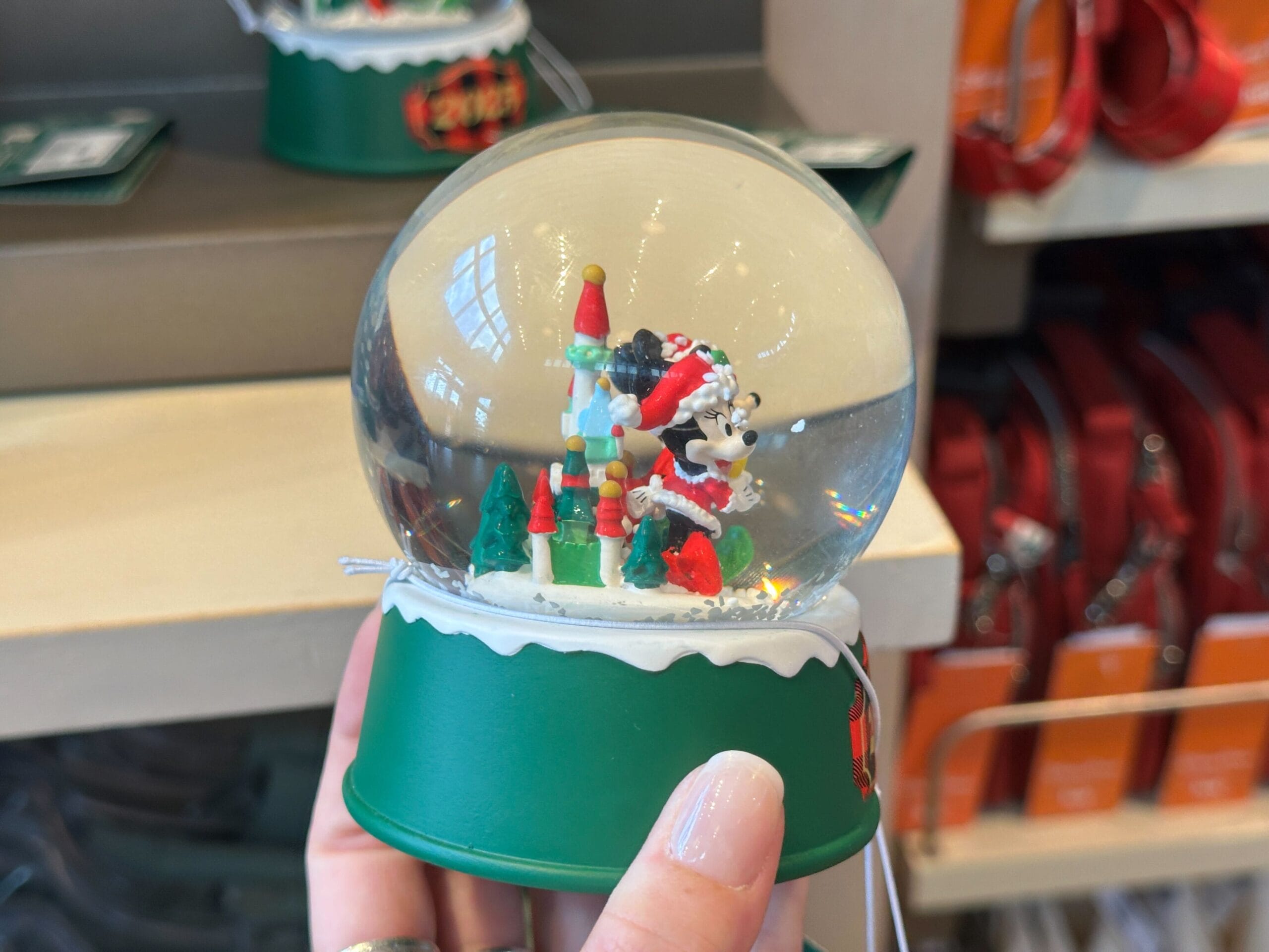A hand displays a green Disney Park snow globe with Minnie Mouse in a Santa hat amid Christmas trees and gifts, no castle visible.
