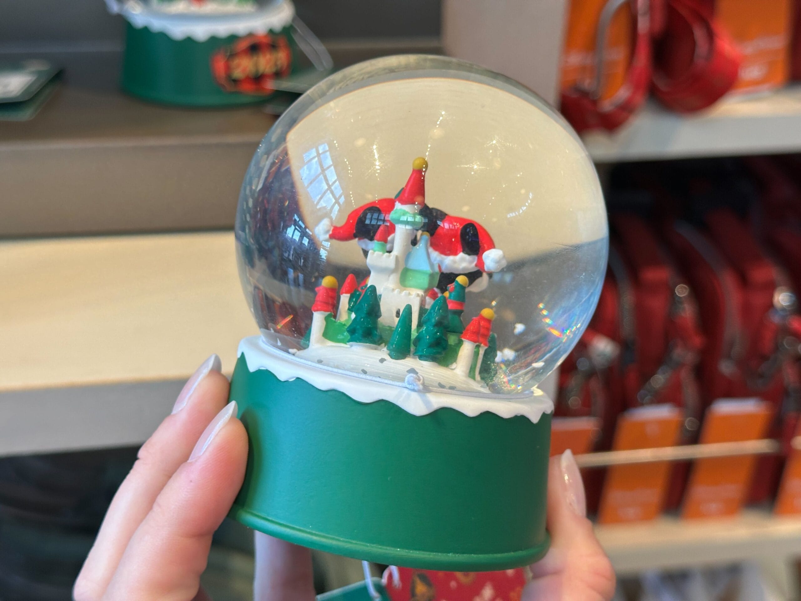 A hand holds a snow globe with a festive holiday house and trees, displayed in a Disney park shop, evoking Main Street’s charm.