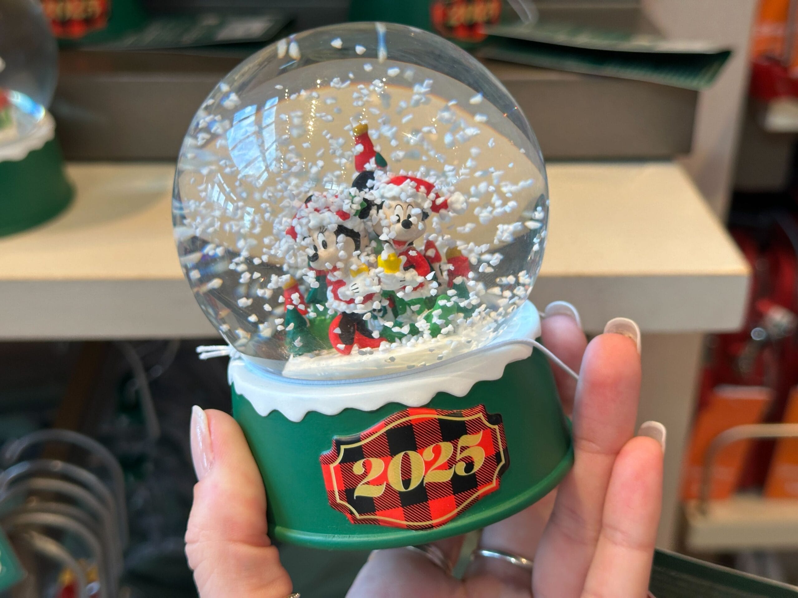 Hand holds a Disney Parks 2023 snow globe with Mickey and Minnie inside, green base with red plaid, reminiscent of park holiday merch.
