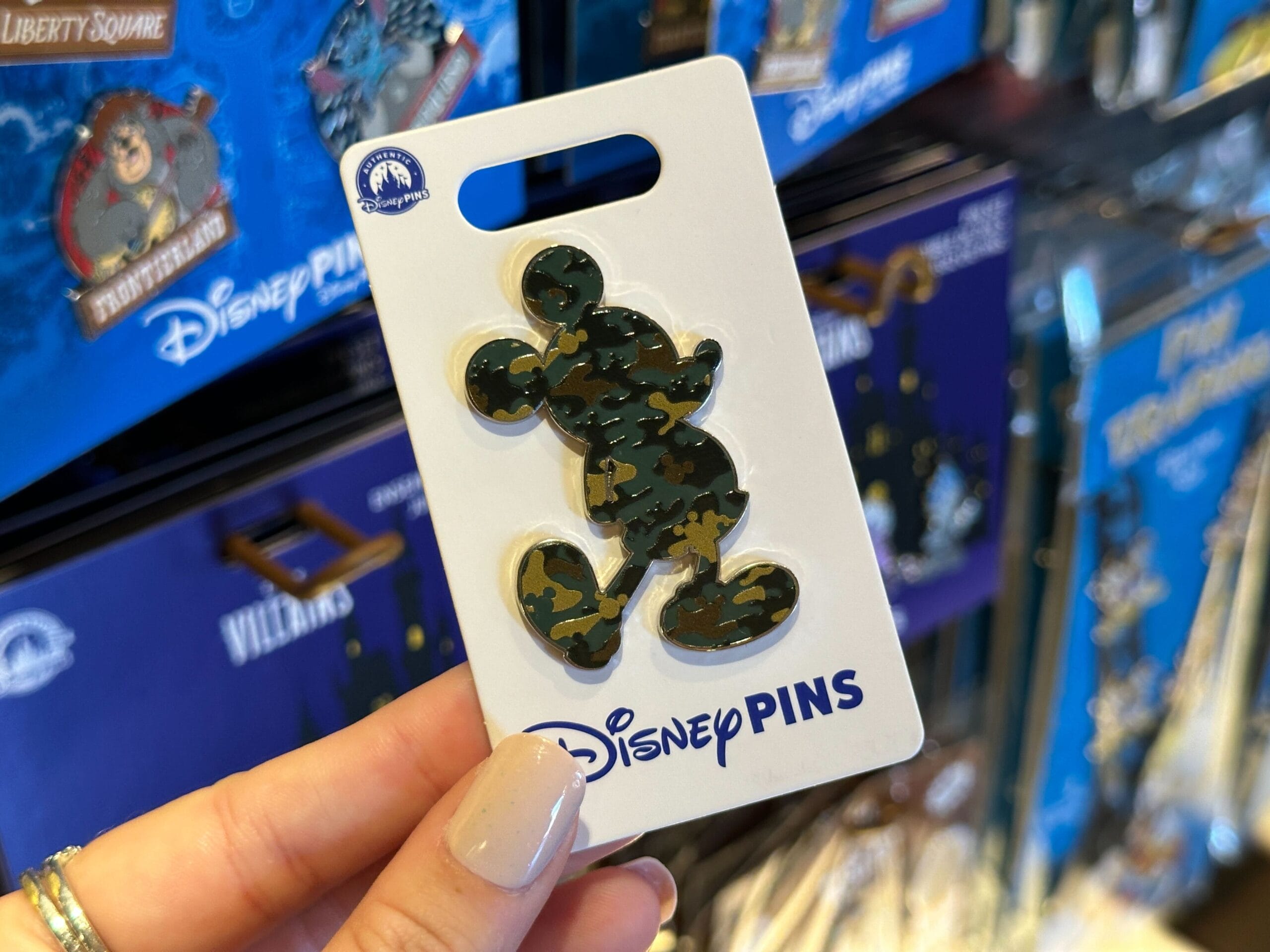 A hand displays a Military Mickey Disney pin in green camo at a Walt Disney World shop, not far from Cinderella Castle—unique to WDW.