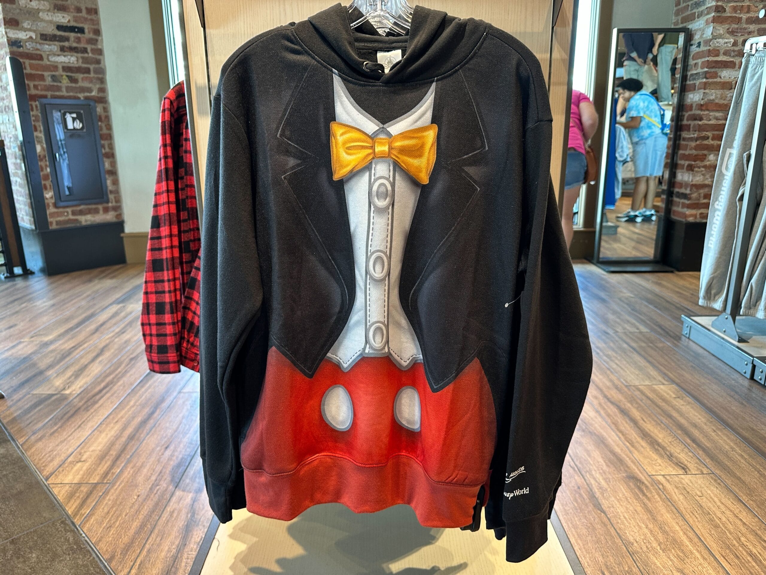 Mickey Mouse tuxedo-style black hoodie with yellow bowtie and red pants details, displayed in a Disney park merchandise shop.