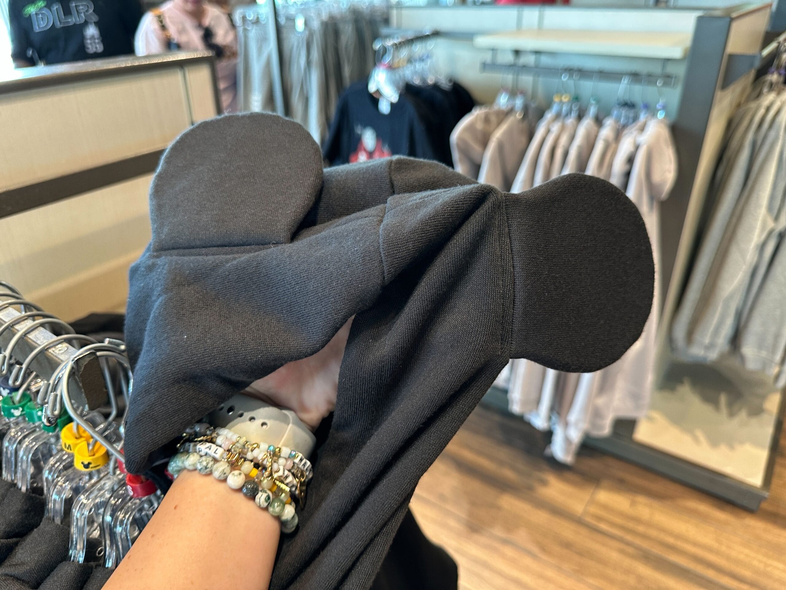 A guest shops for a black hoodie with Mickey Mouse ears in a Disney park store, surrounded by colorful racks of themed apparel.