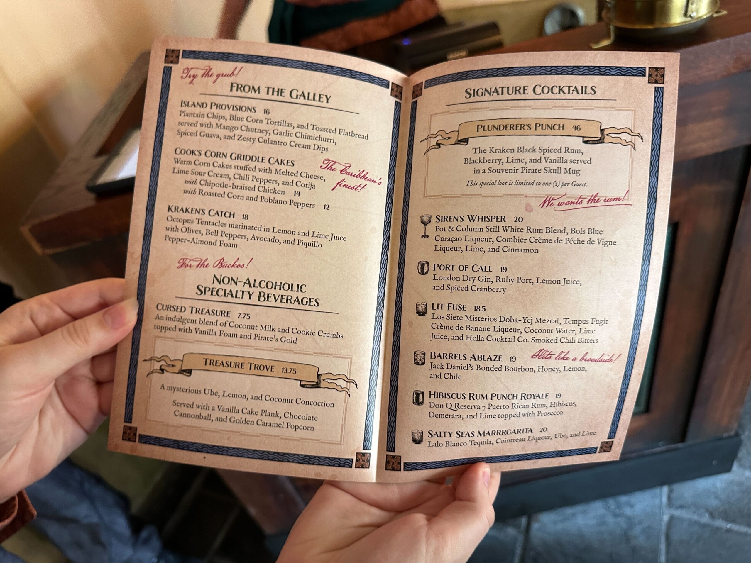 A guest at a Disney park holds a pirate-themed menu featuring food, mocktails, and creative cocktails, reminiscent of park adventures.
