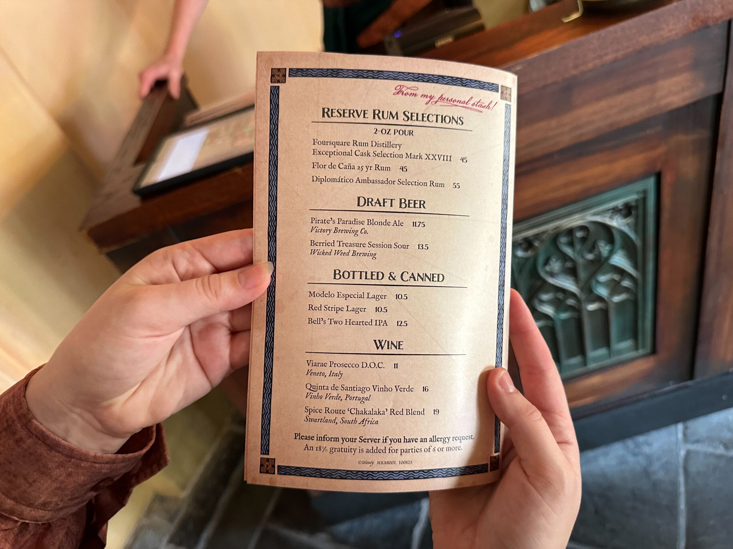 Two hands hold a drinks menu with rum, beer, and wine above a wooden counter, similar to cozy lounges at Disney parks worldwide.