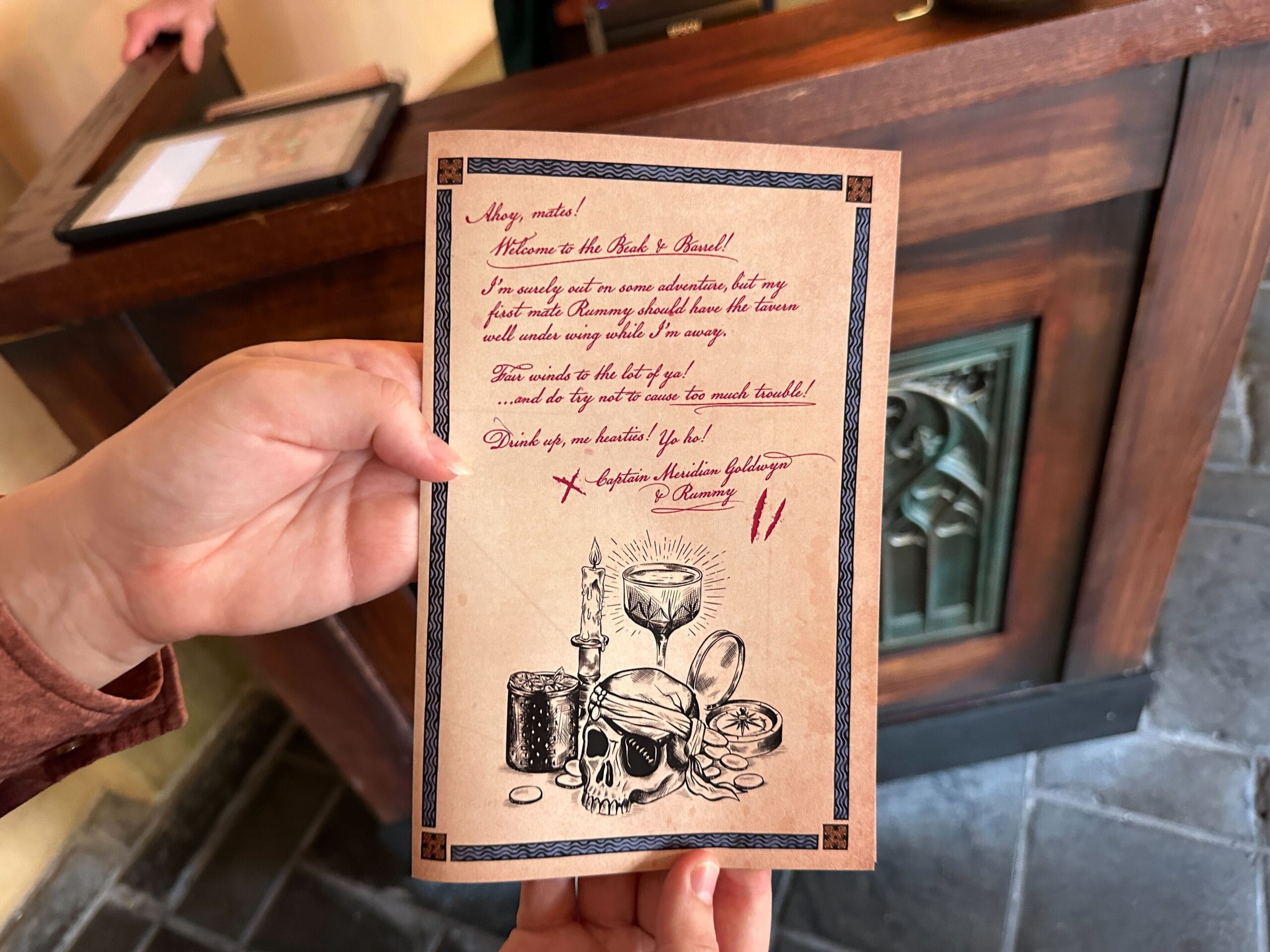 A hand holds a pirate-themed card with skull, candle, and goblet artwork at a wooden counter, evoking Disney’s Pirates attractions.