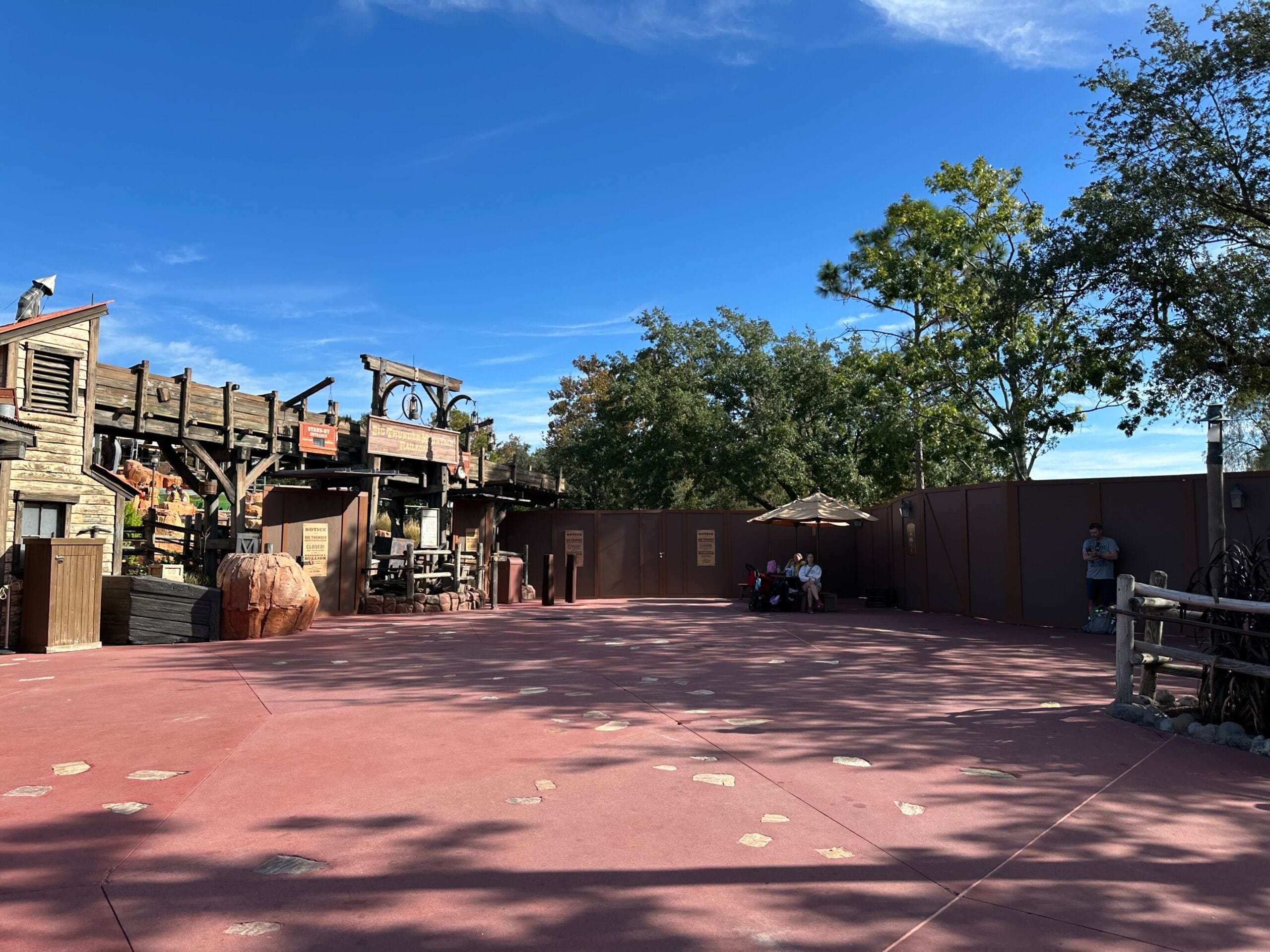 A quiet courtyard in a Disney park with construction walls, themed wooden buildings, and guests relaxing under umbrellas. No castle visible.