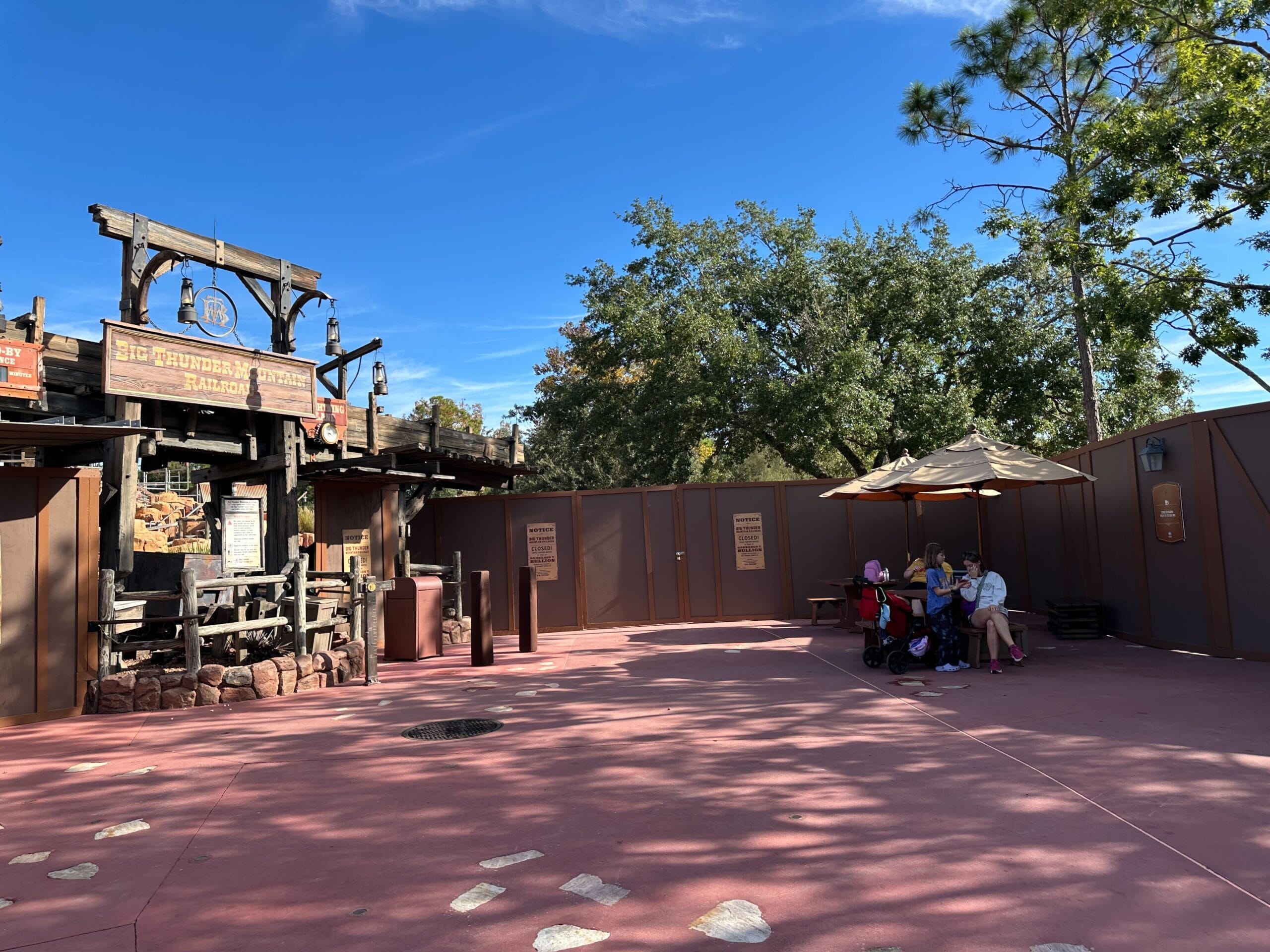 A family relaxes in the shade of an umbrella beside construction walls at Big Thunder Mountain Railroad's entrance on a sunny Disney day.