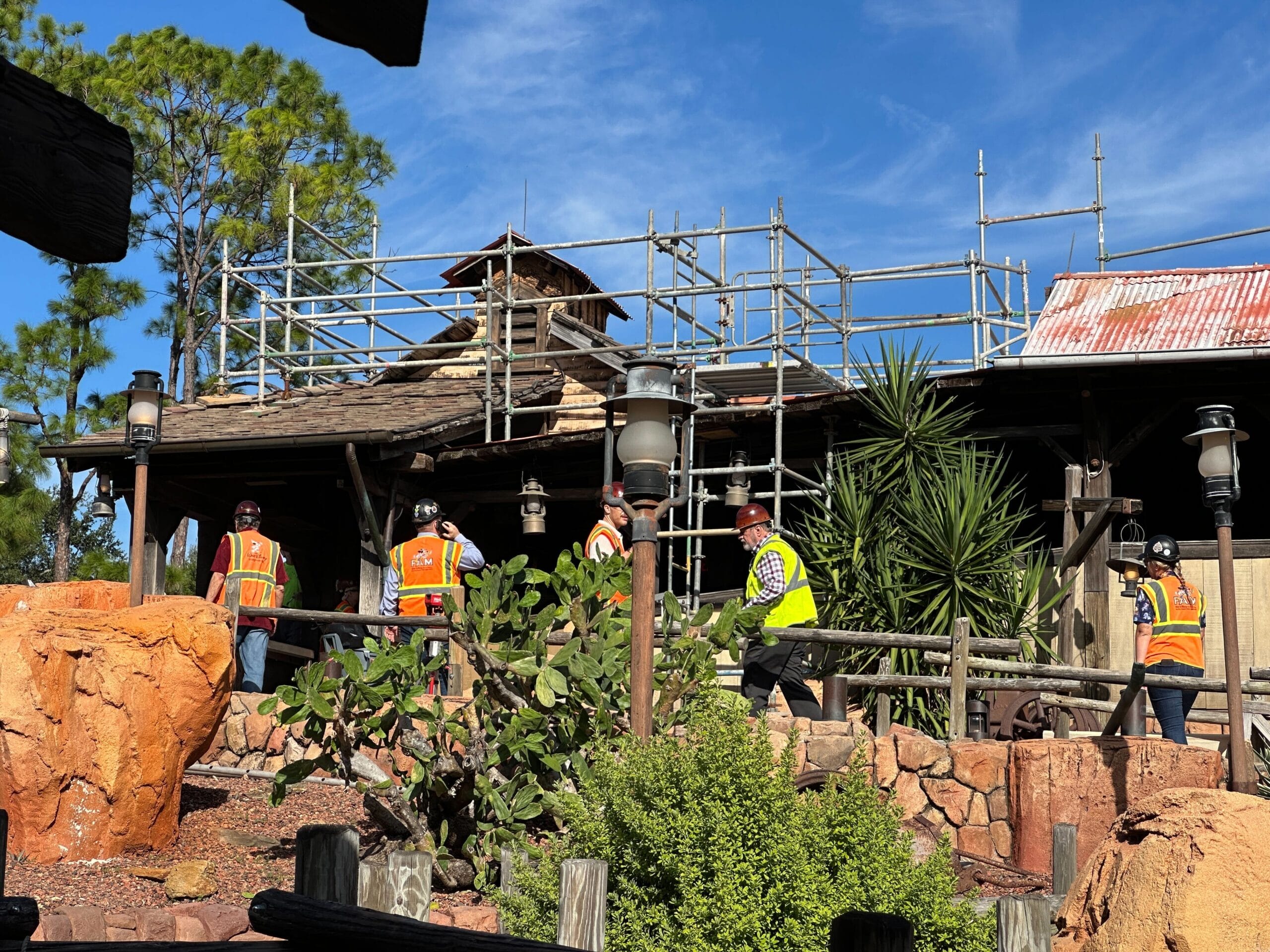 Construction workers in safety gear walk near scaffolding by a rustic building, reminiscent of Disney park architecture, with lush landscaping.