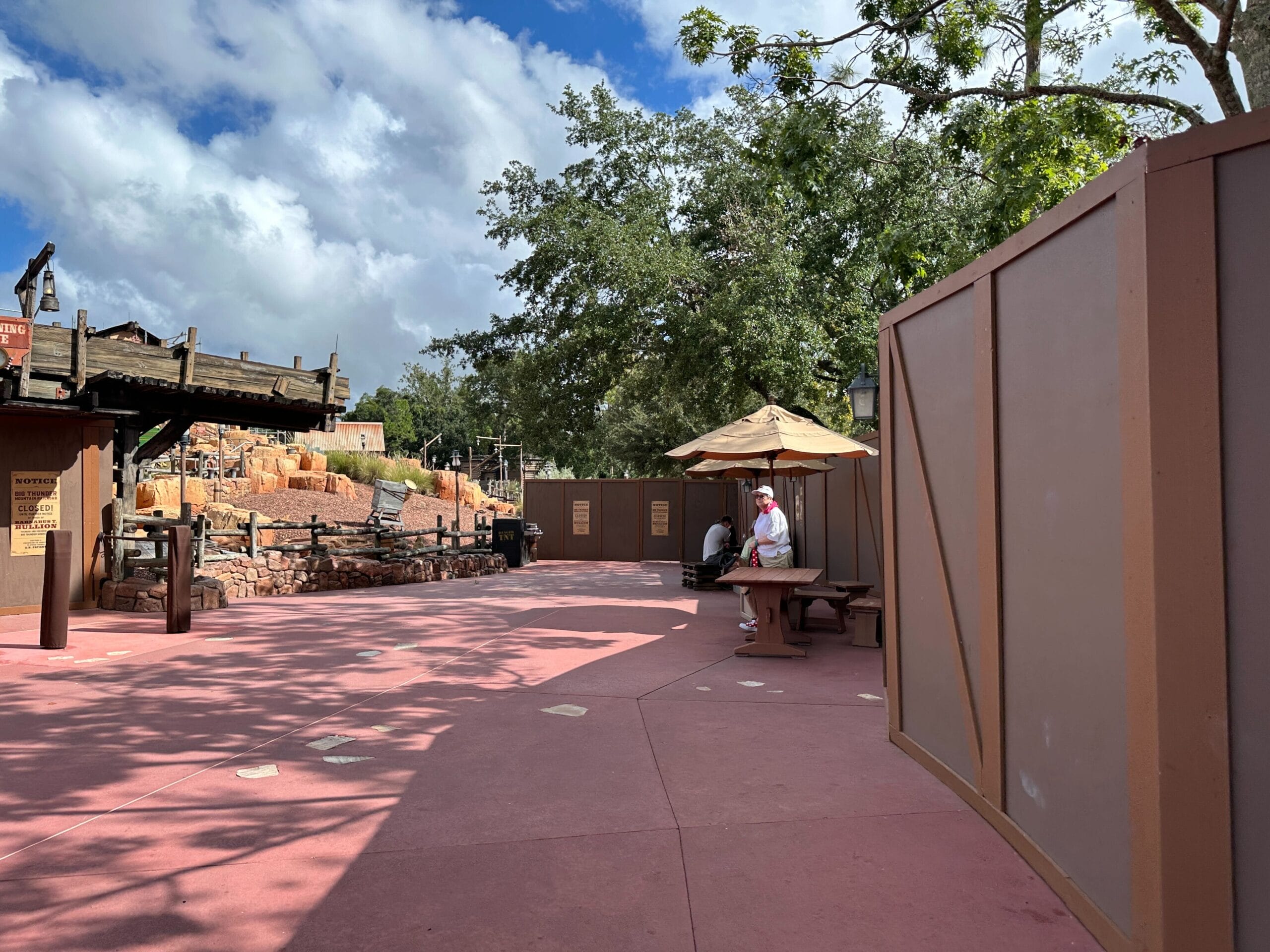 Near Magic Kingdom, a quiet walkway lines up next to construction walls, with a Cast Member under an umbrella and trees in back.