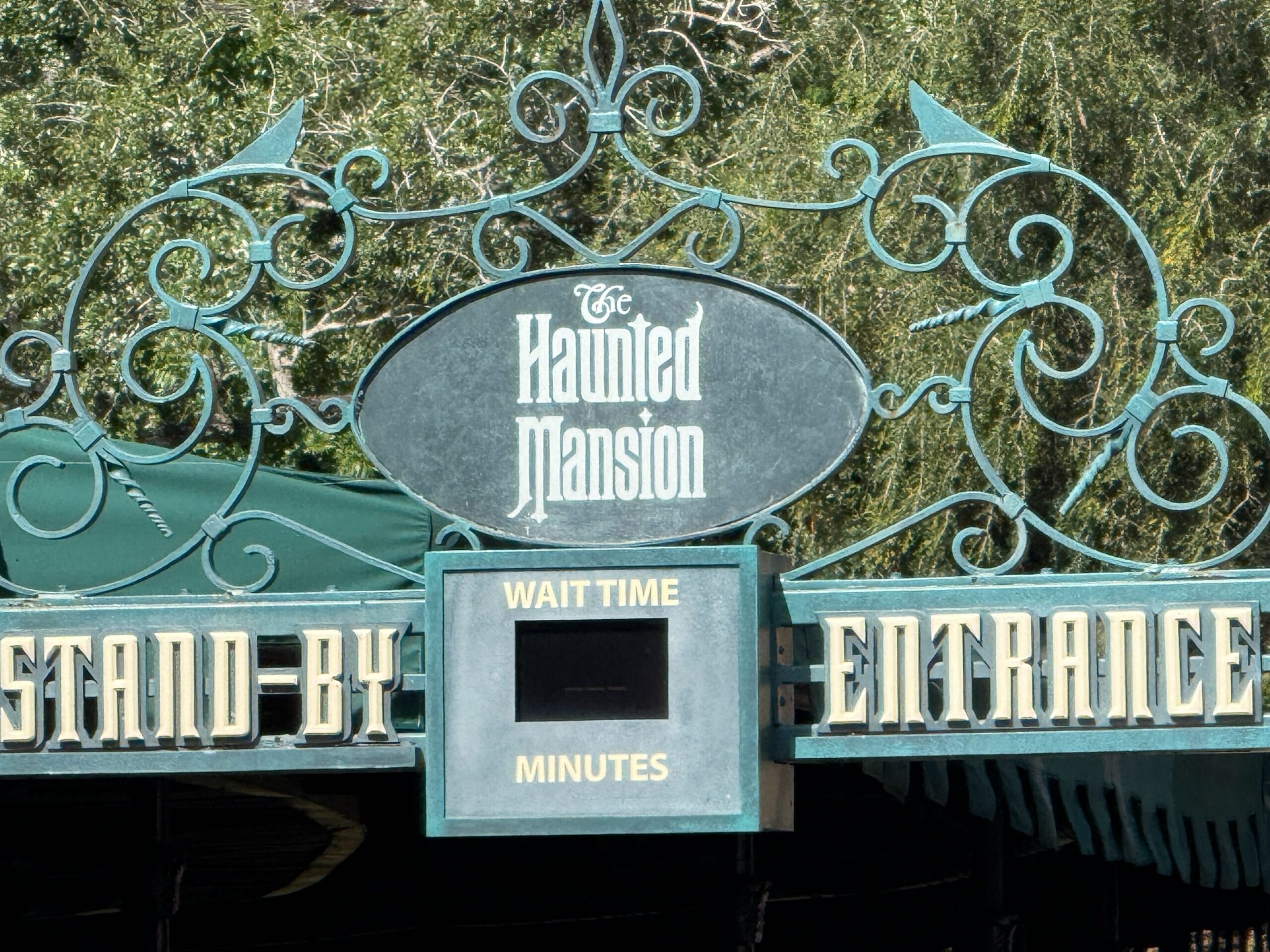 Detailed Haunted Mansion entrance sign at Disney park, wait time blank, lush greenery around, evoking classic gothic style.