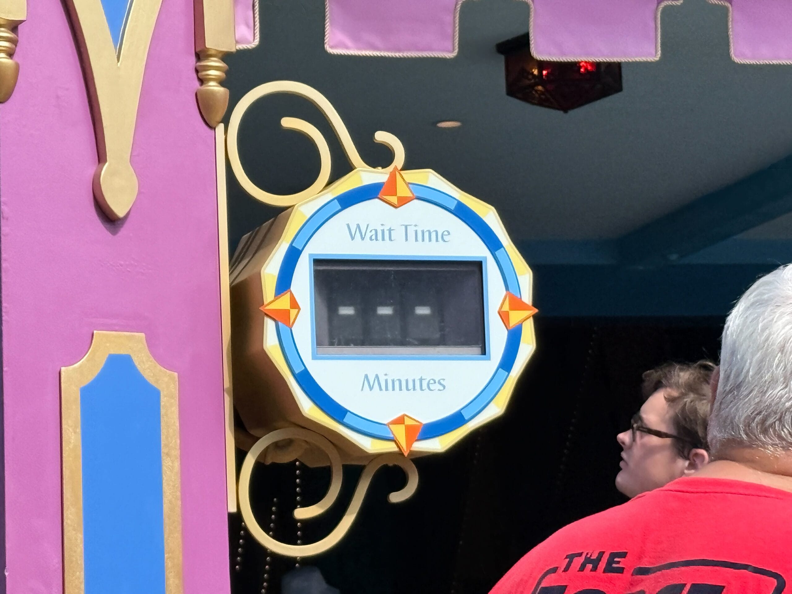 A digital wait time sign at a Disney park displays blank spaces, as nearby guests check for updates under classic themed decor.