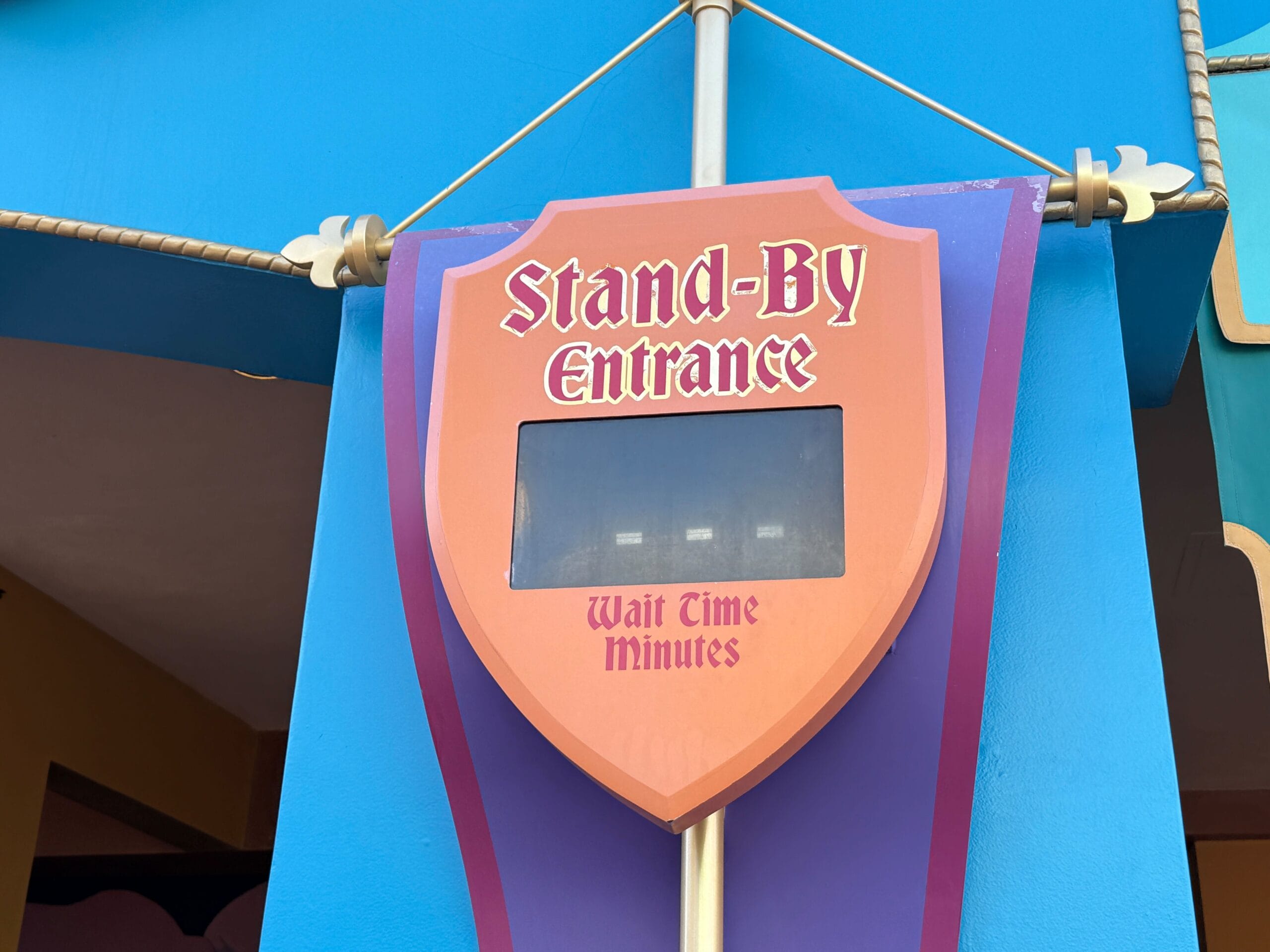 Red shield-shaped "Stand-By Entrance" sign with empty wait time window, colorful Disney Park theming in the background.