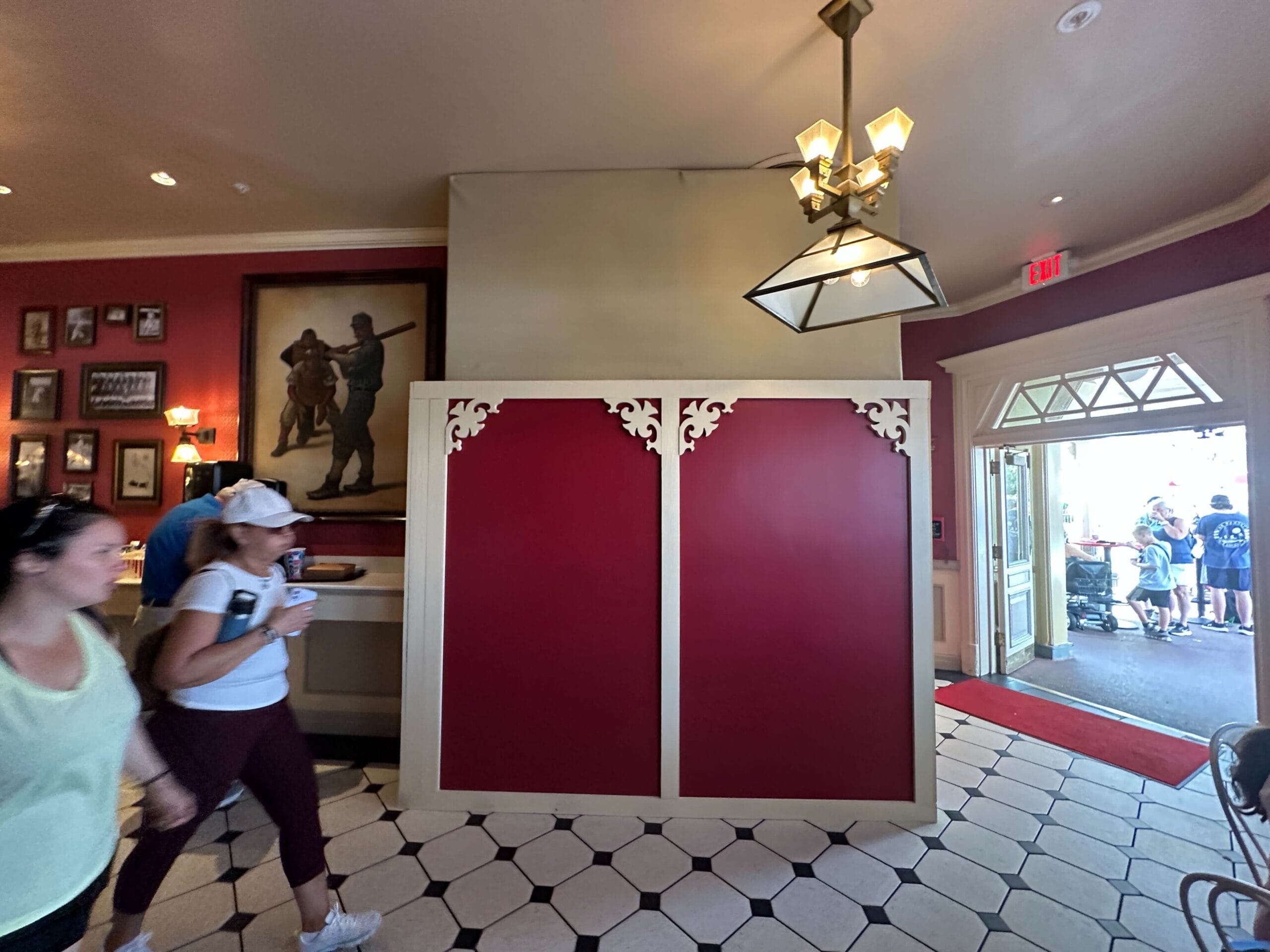 Two women stroll past a red and cream partition in a Disney park lobby, with others visible through an open doorway behind them.