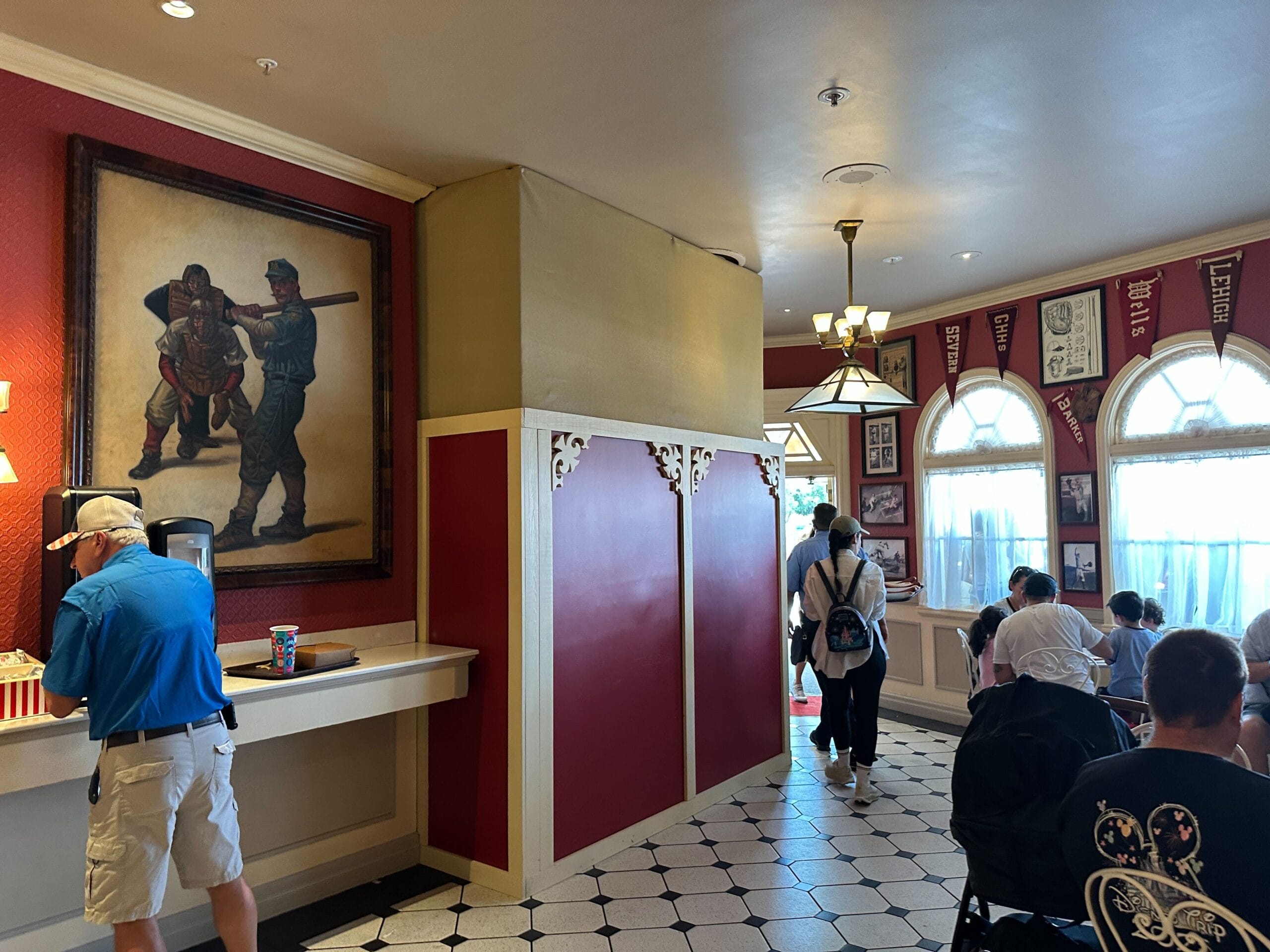 Guests dine inside a Disney park restaurant with red and white decor; a large painting and popcorn machine are on the left wall.