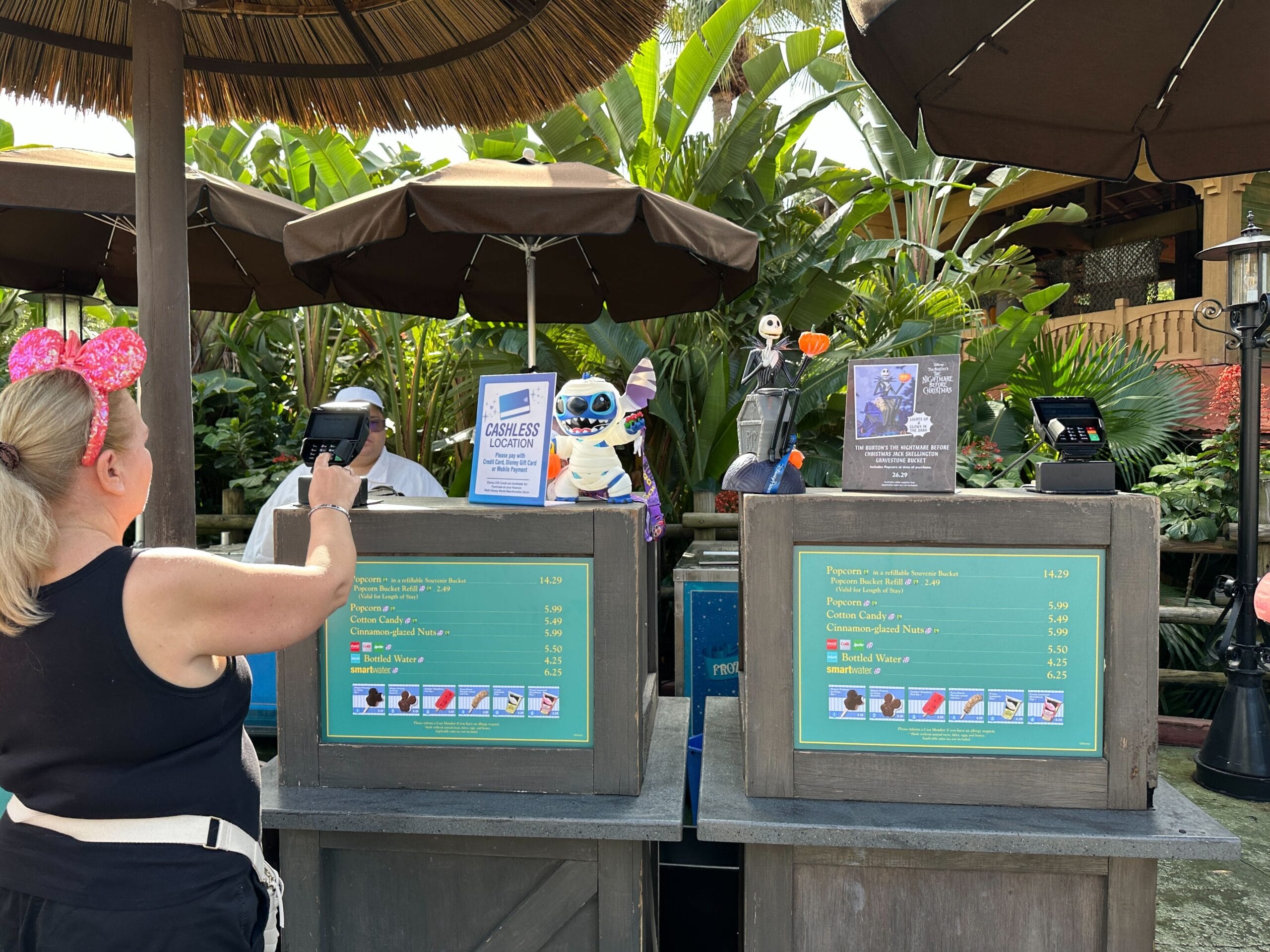 A woman in pink Minnie ears uses cashless payment at a Walt Disney World snack cart featuring Stitch and Jack Skellington toys.