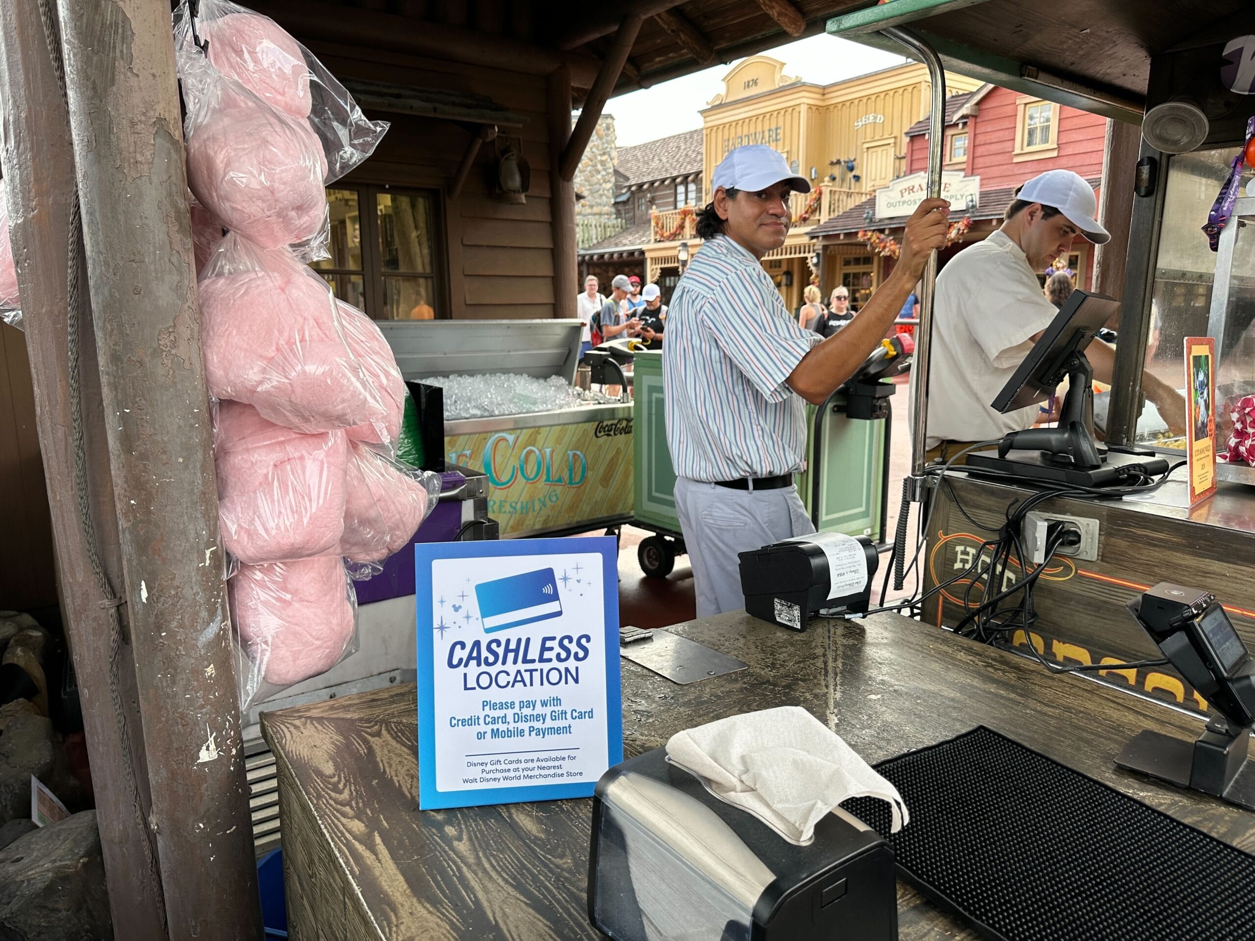 A vendor in a striped shirt stands beside a cashless snack cart at Walt Disney World, with no castle visible in the background.