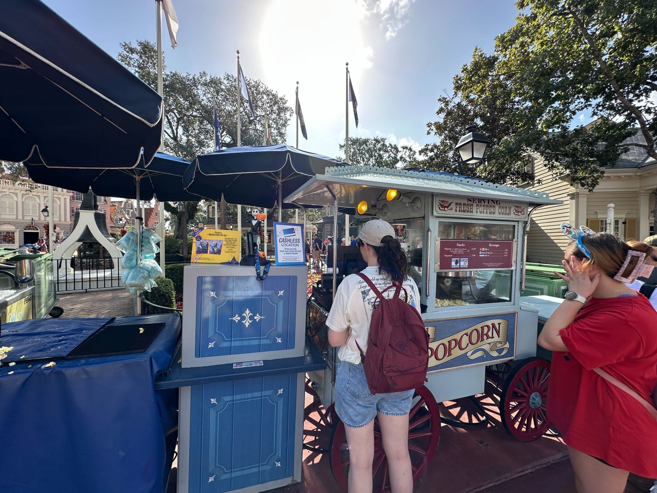 Two guests at a Disney World snack cart bask in sunshine; blue umbrellas and trees evoke classic Magic Kingdom park vibes.