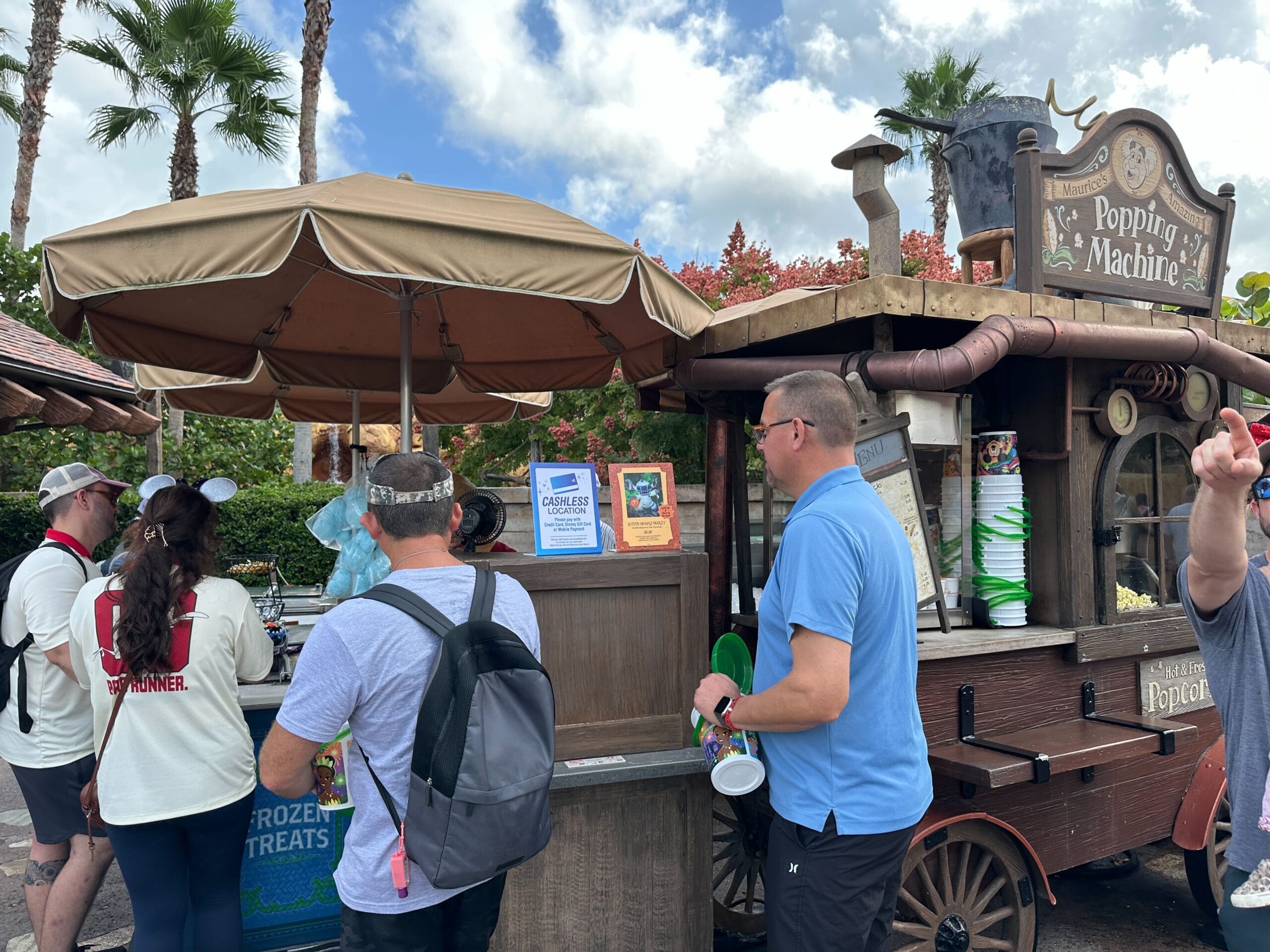 Guests queue for the cashless "Popping Machine" snack cart beneath palm trees in sunny Walt Disney World—no castle in view here.