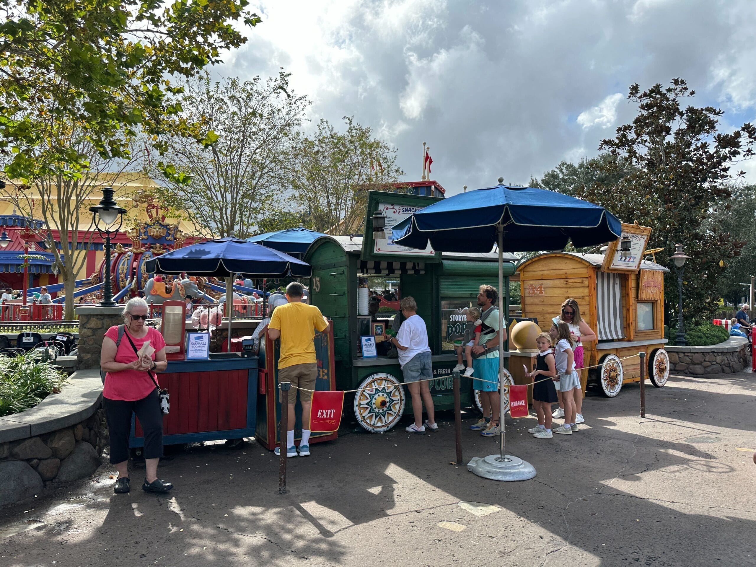 Guests wait in line at a vibrant snack cart with blue umbrellas on a sunny day at Walt Disney World—no castle is visible.