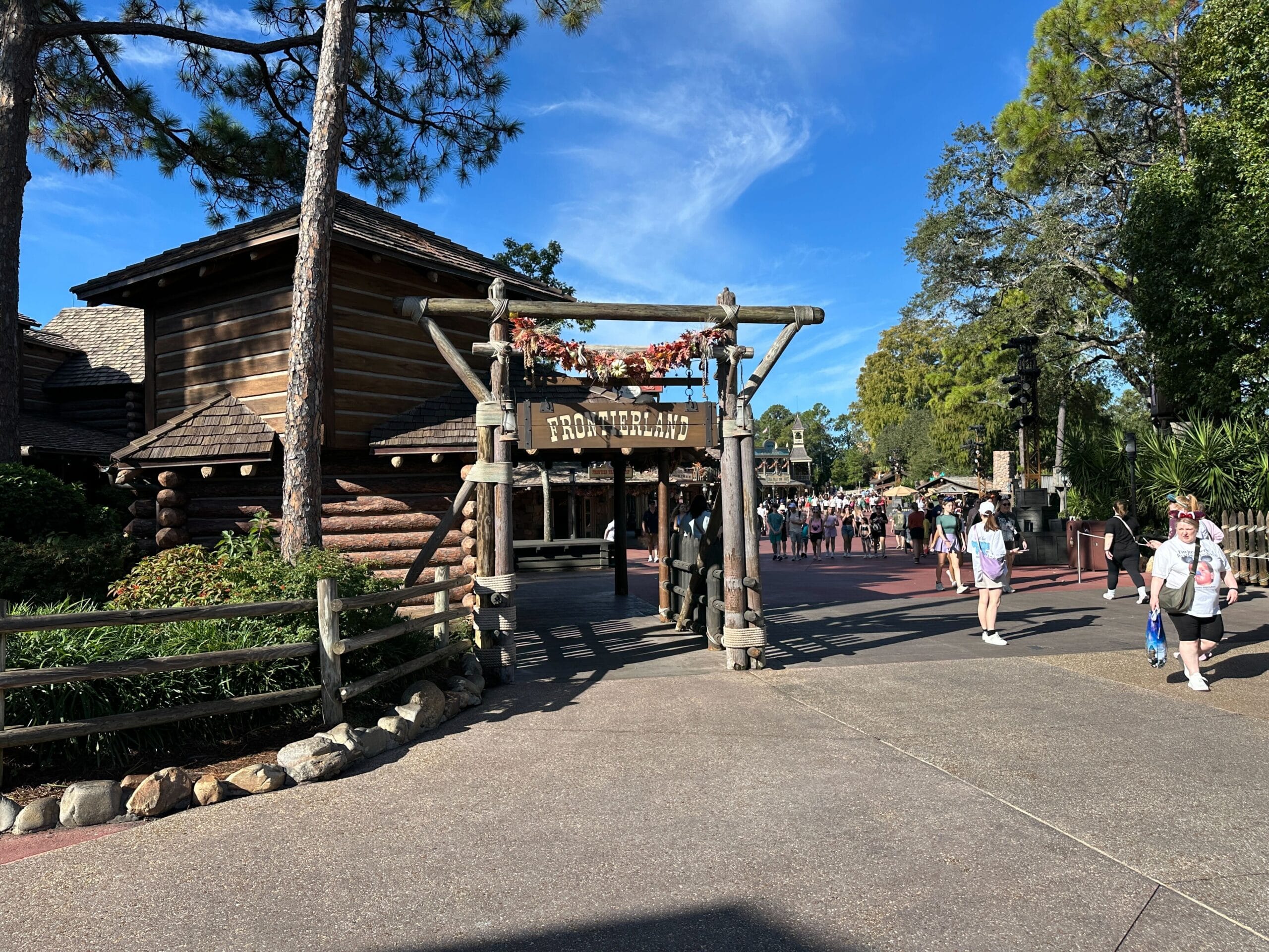 Entrance to Frontierland at a Disney park, with guests strolling by rustic log-style buildings and trees; no castle in view here.