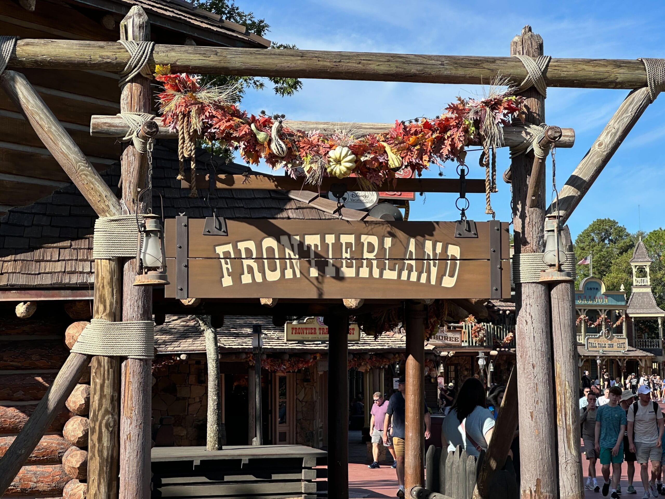 Frontierland sign hanging under a fall leaf arch at a Disney park, people strolling below; no castle visible in this scene.