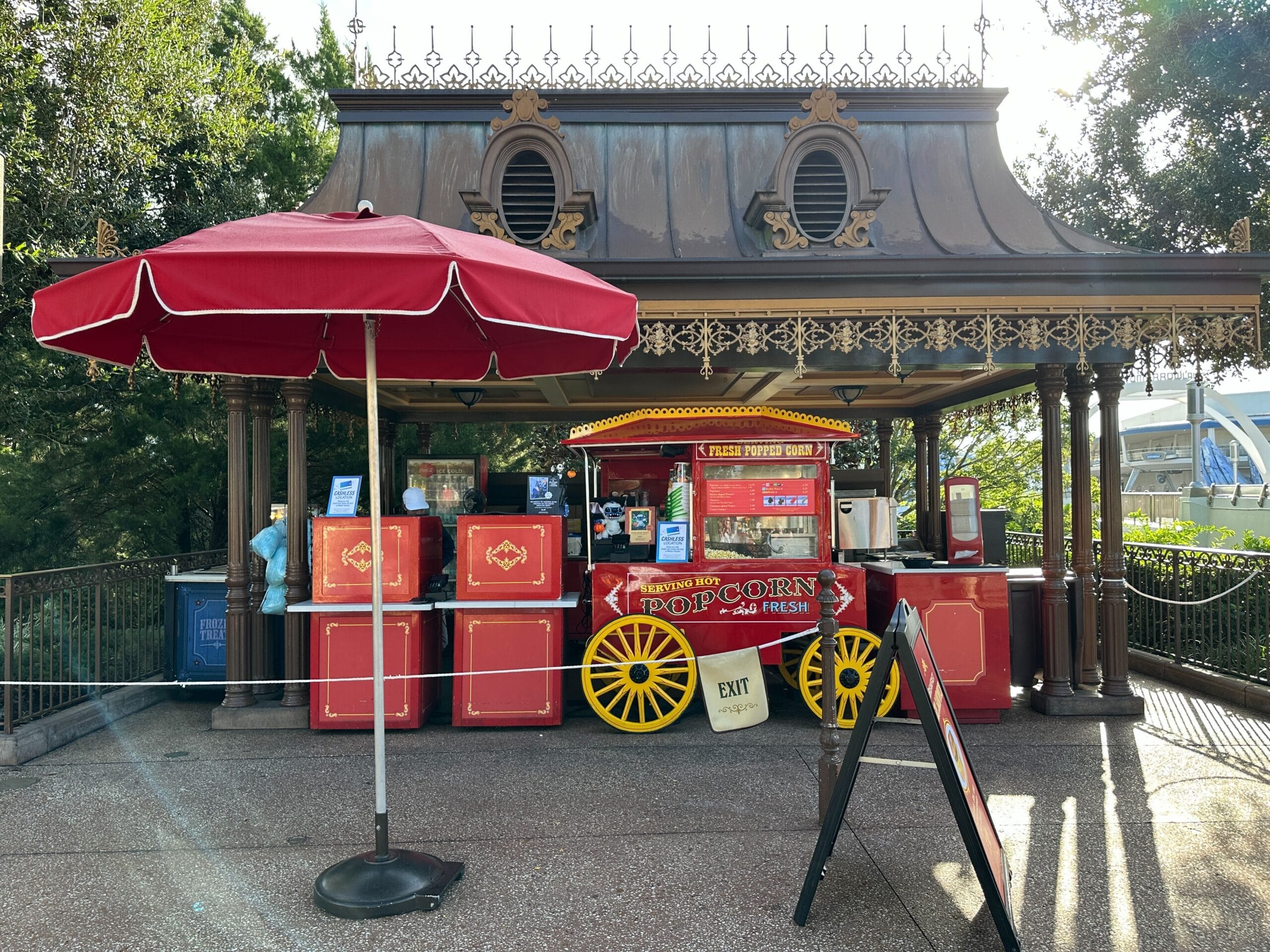 A classic red popcorn cart with a yellow wheel sits under a pavilion in a Disney park, near a red umbrella and trees on a sunny day.