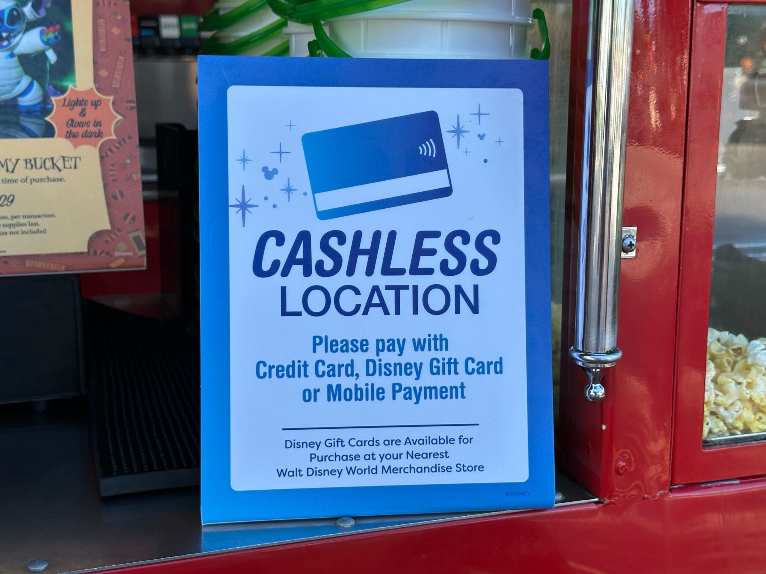 At a Disney park concession stand, a sign reads: "Cashless Location—please pay with card or mobile payment."