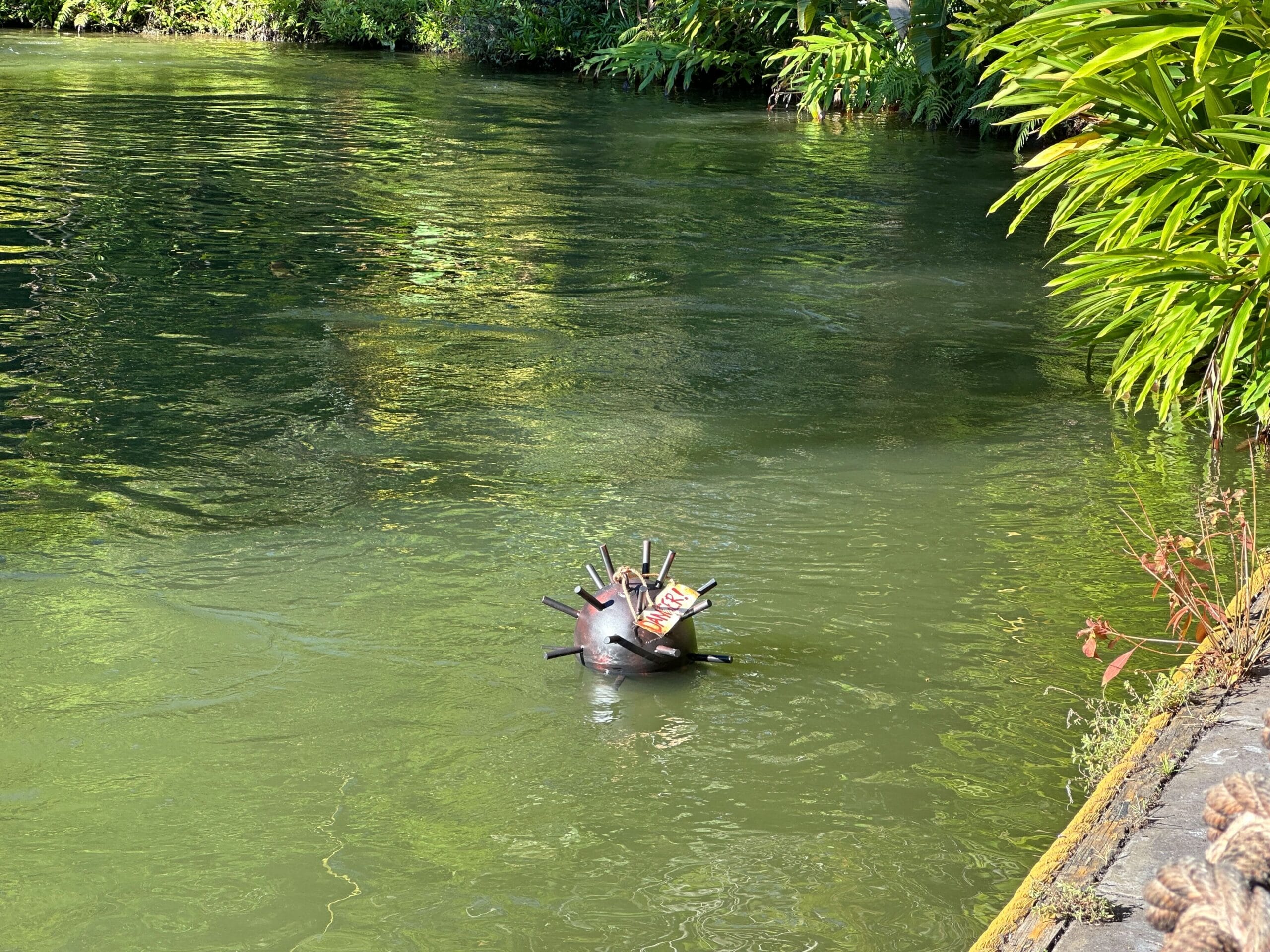 A floating metal sea mine replica in a green pond at a Disney park, surrounded by lush landscaping and themed stone edging.