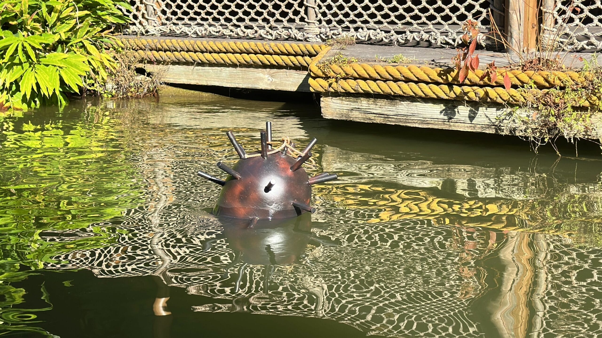 A floating naval mine with spikes sits in greenish water near a dock at a Disney park, surrounded by rope and themed greenery.