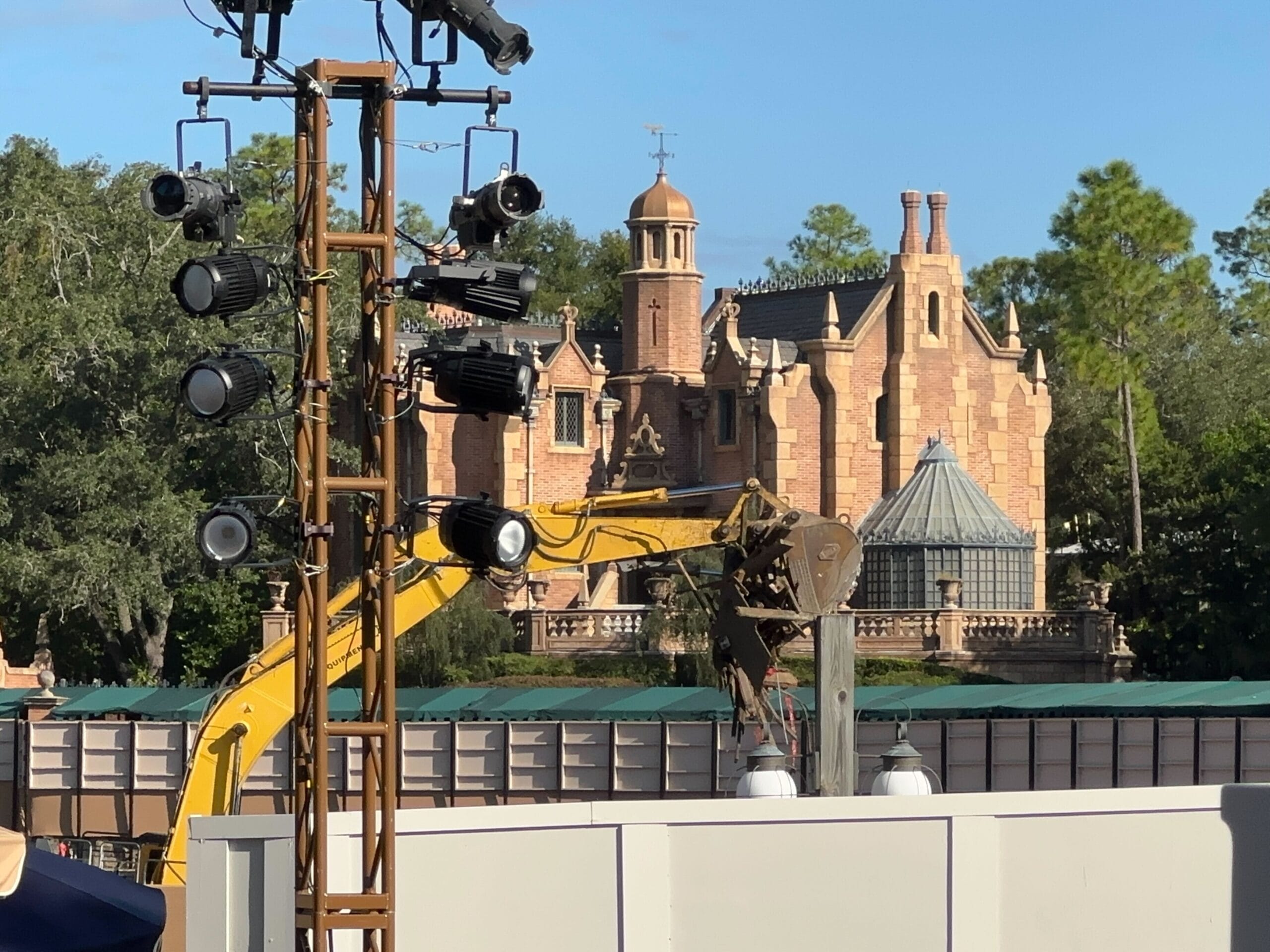 A yellow excavator and stage lights by the Haunted Mansion with trees and barriers in Magic Kingdom.