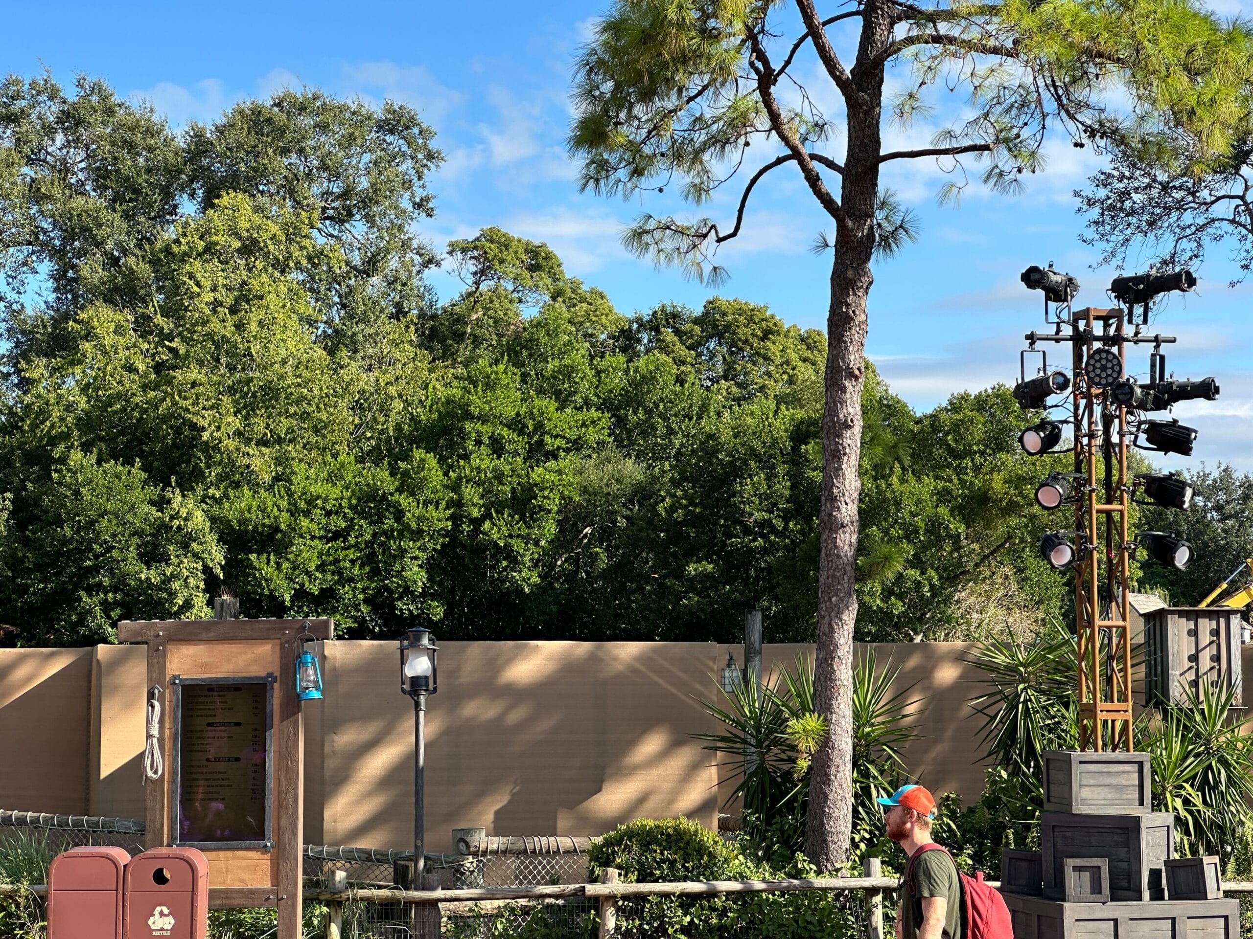 A guest in a red hat strolls past tan construction walls, trees, and stage lights beneath a clear blue sky at a Disney park.