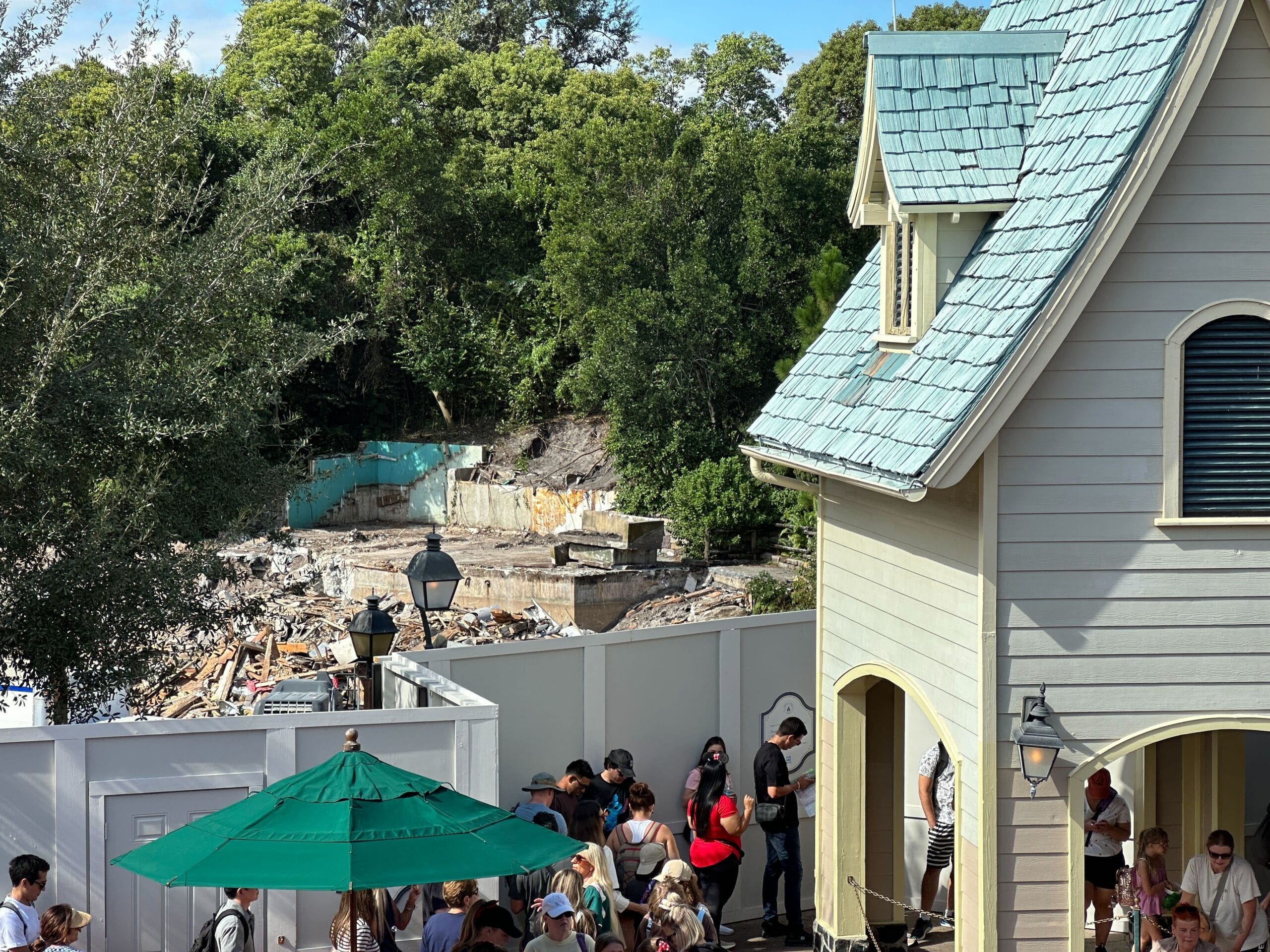 A crowd waits by a pale house near a white fence, where trees surround the Aunt Polly's demolition site.